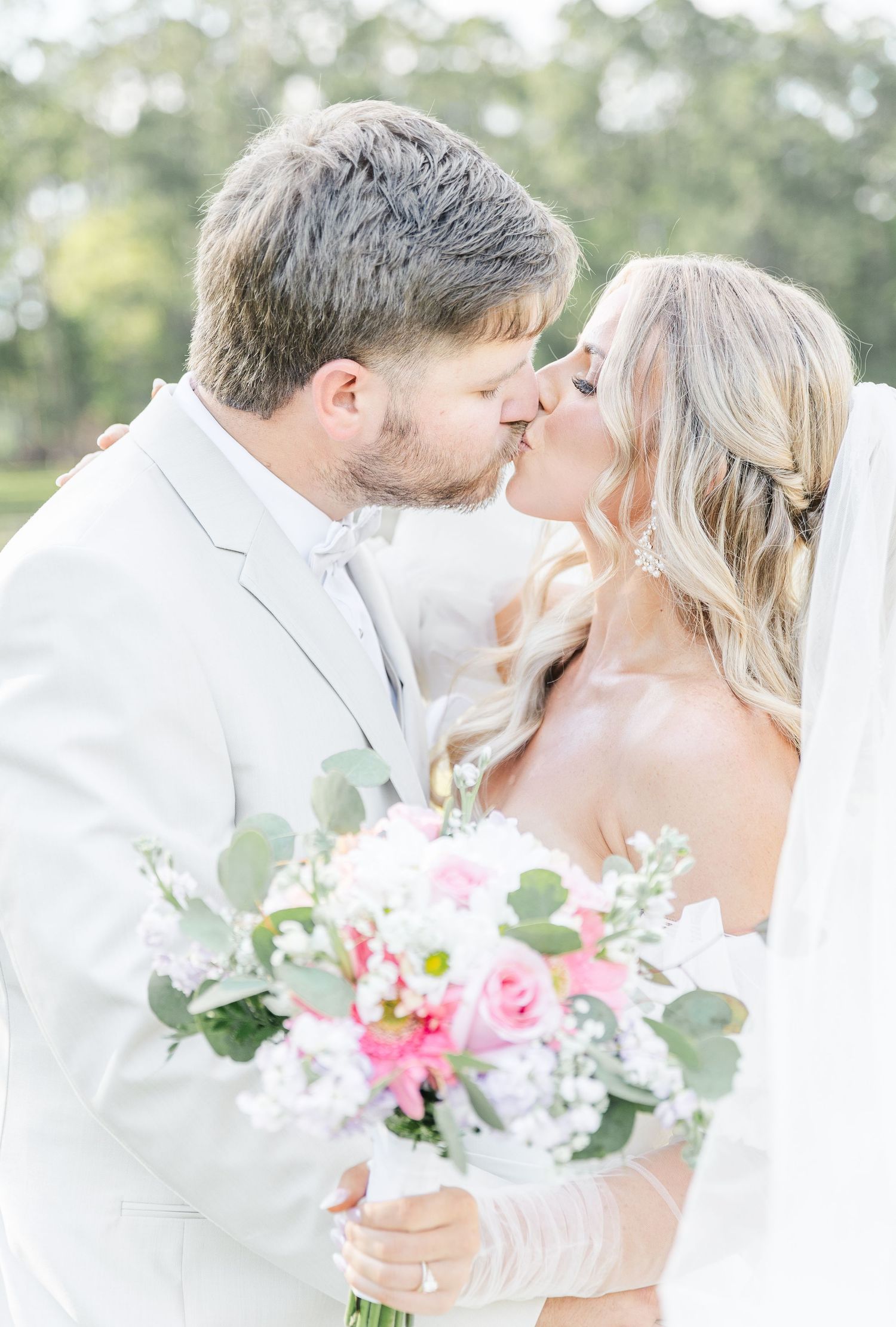 Romantic wedding portrait of newlyweds sharing a tender kiss while holding a pink and white floral bouquet.