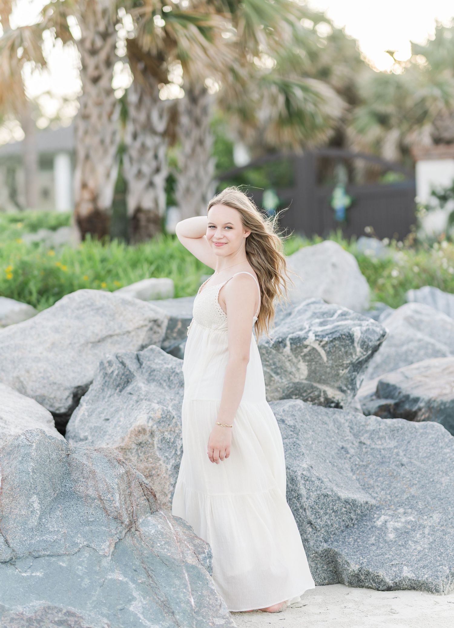 A white maxi dress flows in the breeze as someone poses among coastal rocks and palm trees.