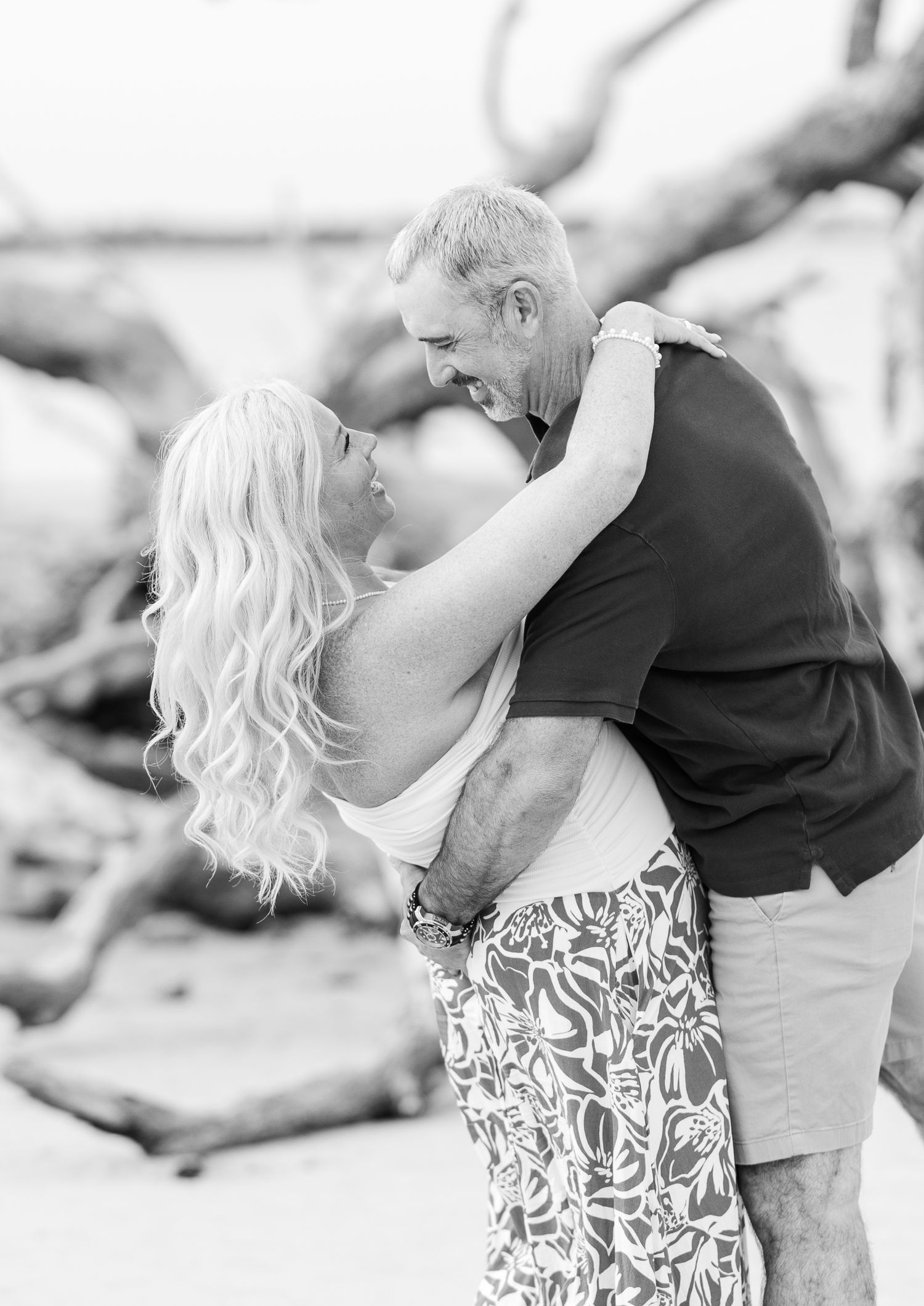 A romantic black and white photo of a couple embracing on a beach with driftwood in the background.