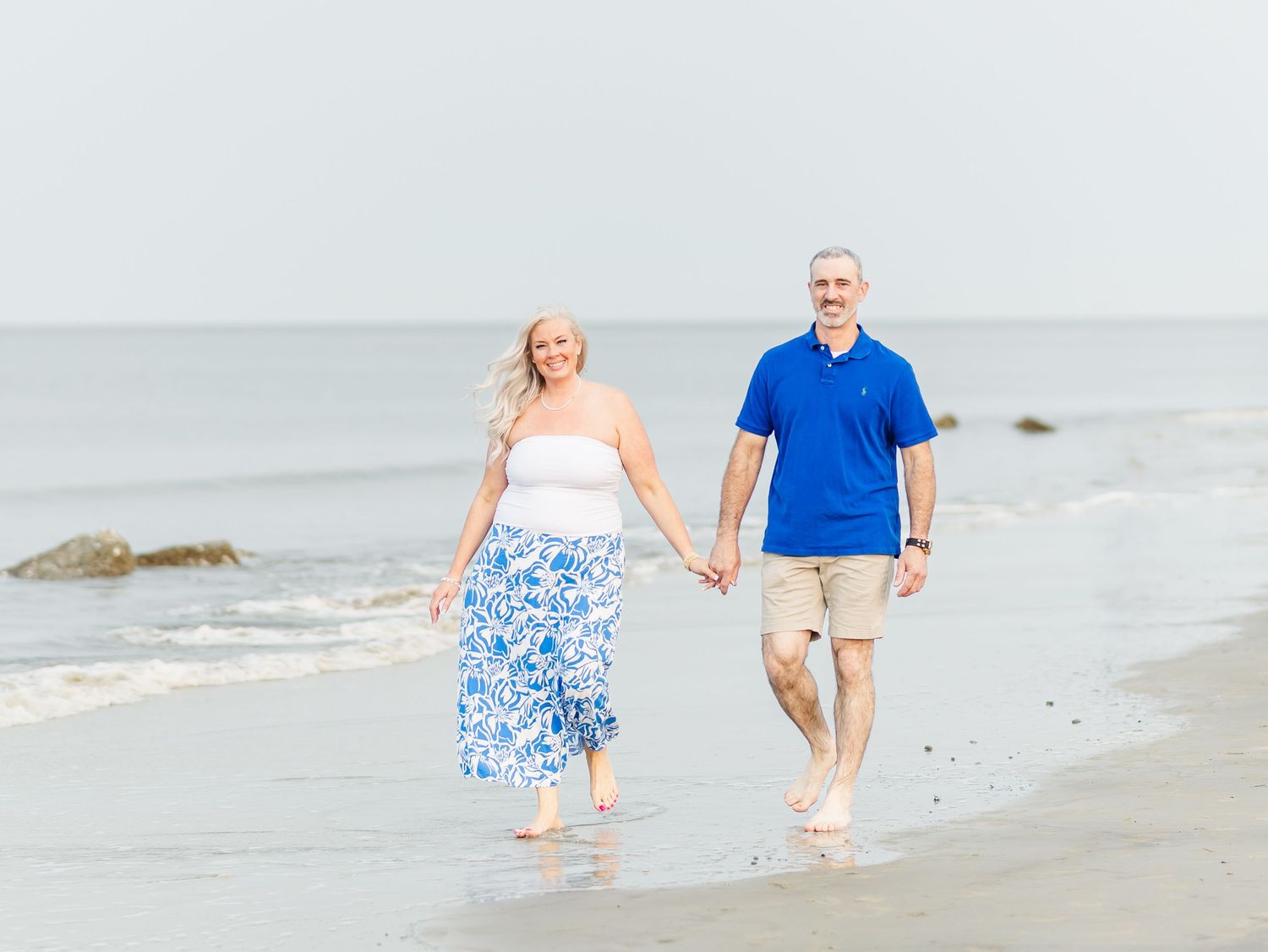 A couple wearing coordinating blue and white outfits strolls along a sandy beach at the ocean's edge.