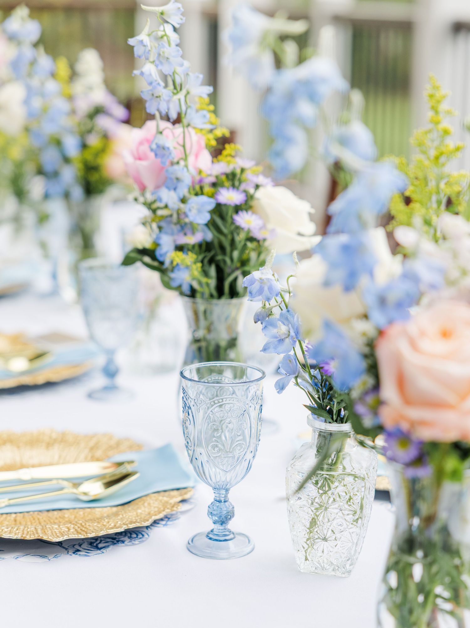 Elegant wedding tablescape with blue goblets, gold flatware, and pastel floral arrangements featuring blue delphiniums and pink roses.