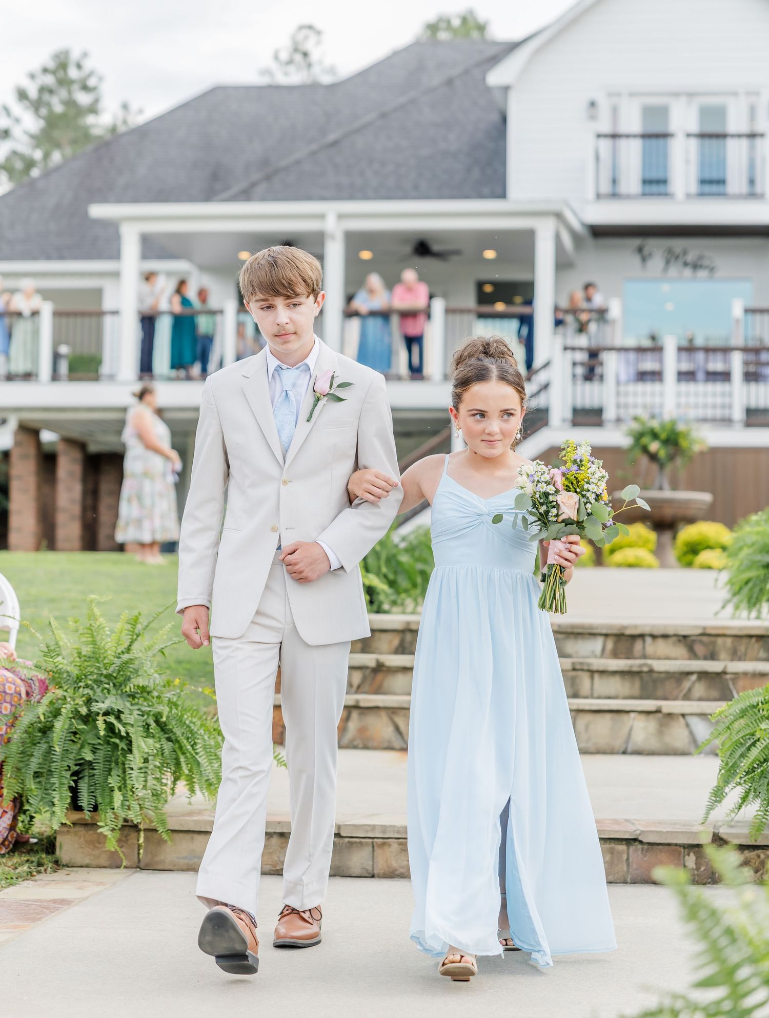 A young usher in a gray suit escorts a junior bridesmaid in a light blue gown down the aisle.