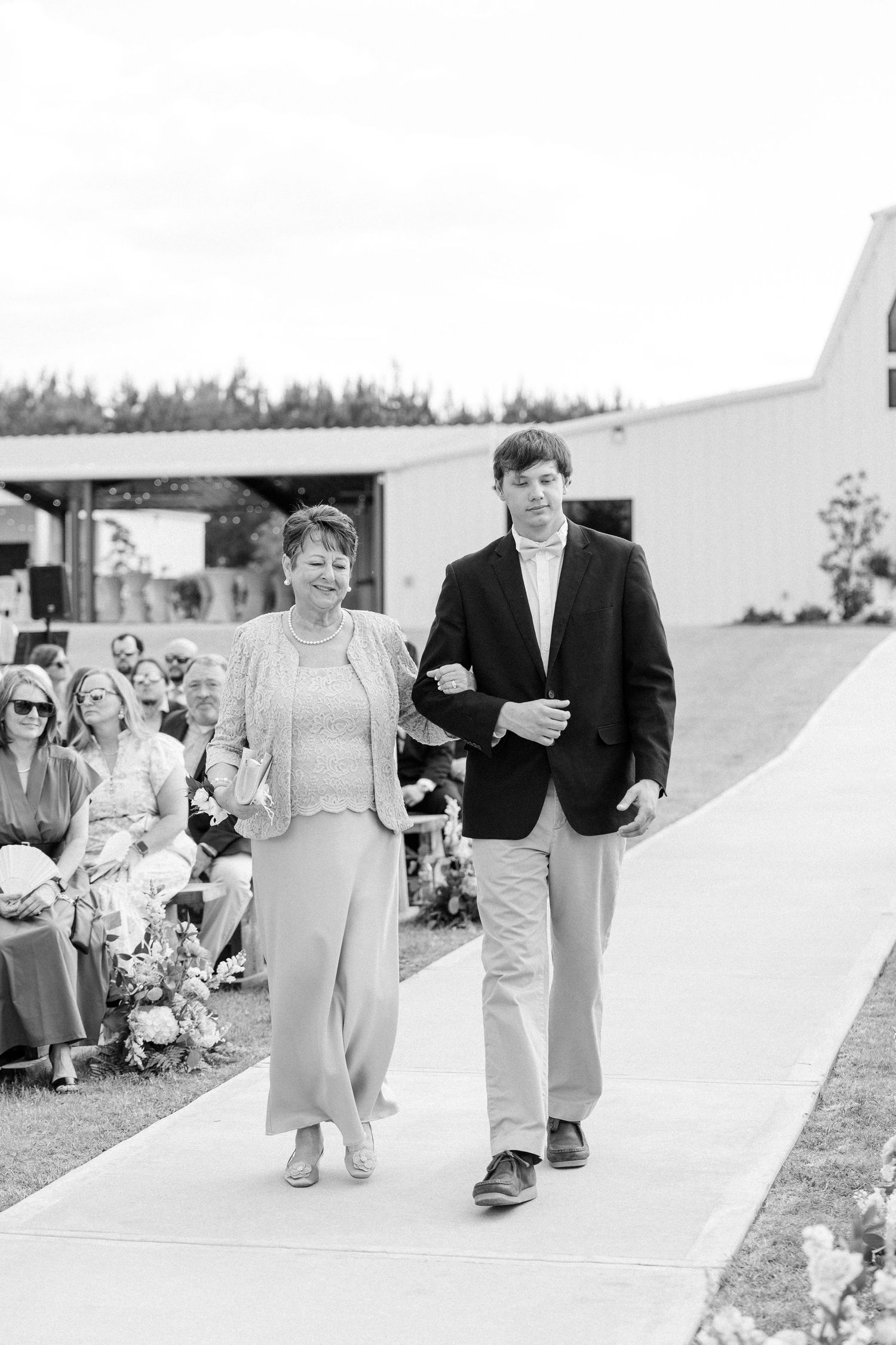 Black and white photo of woman and young man walking down outdoor wedding ceremony aisle.