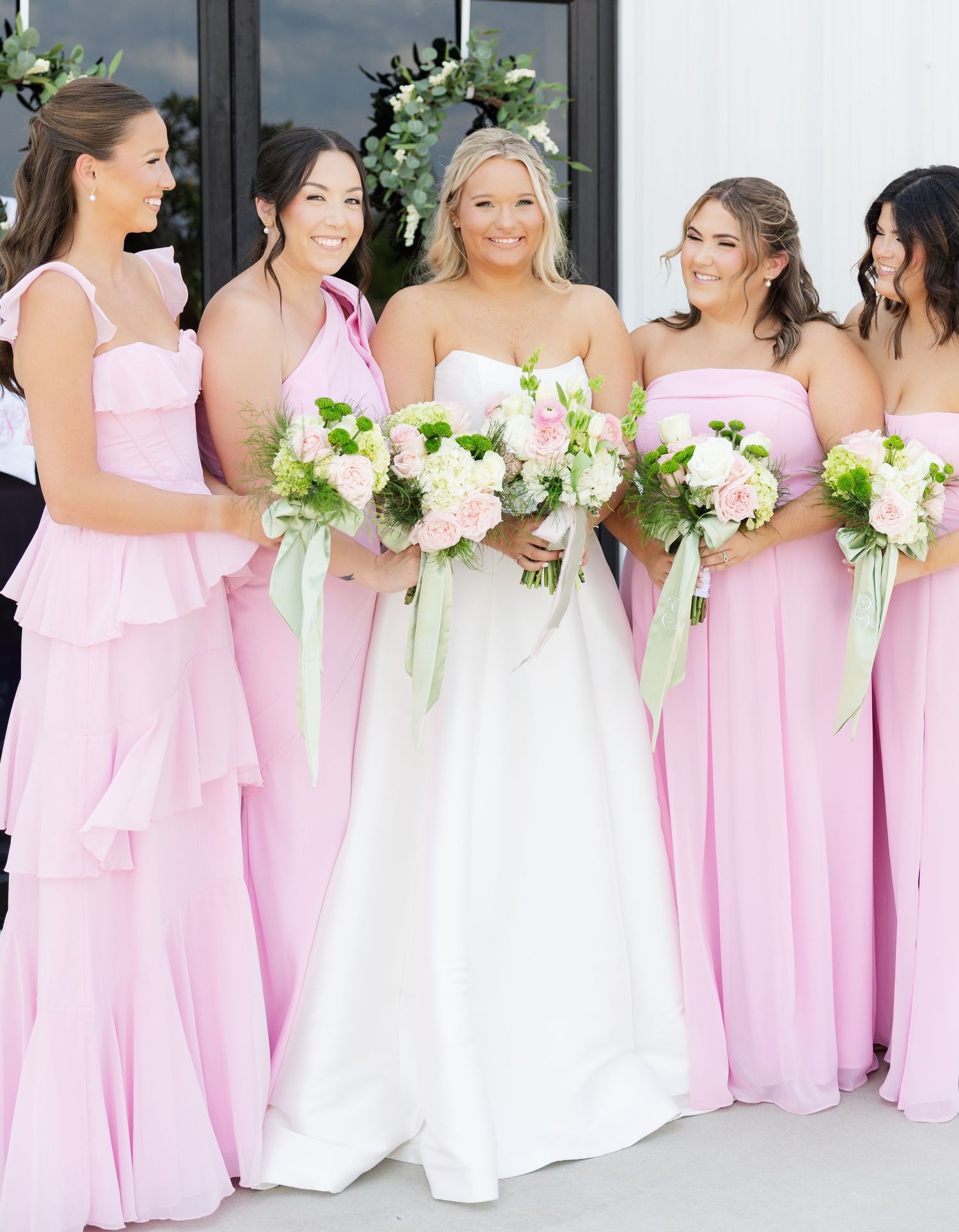 Bride in white gown with bridesmaids in pink dresses holding floral bouquets outdoors.