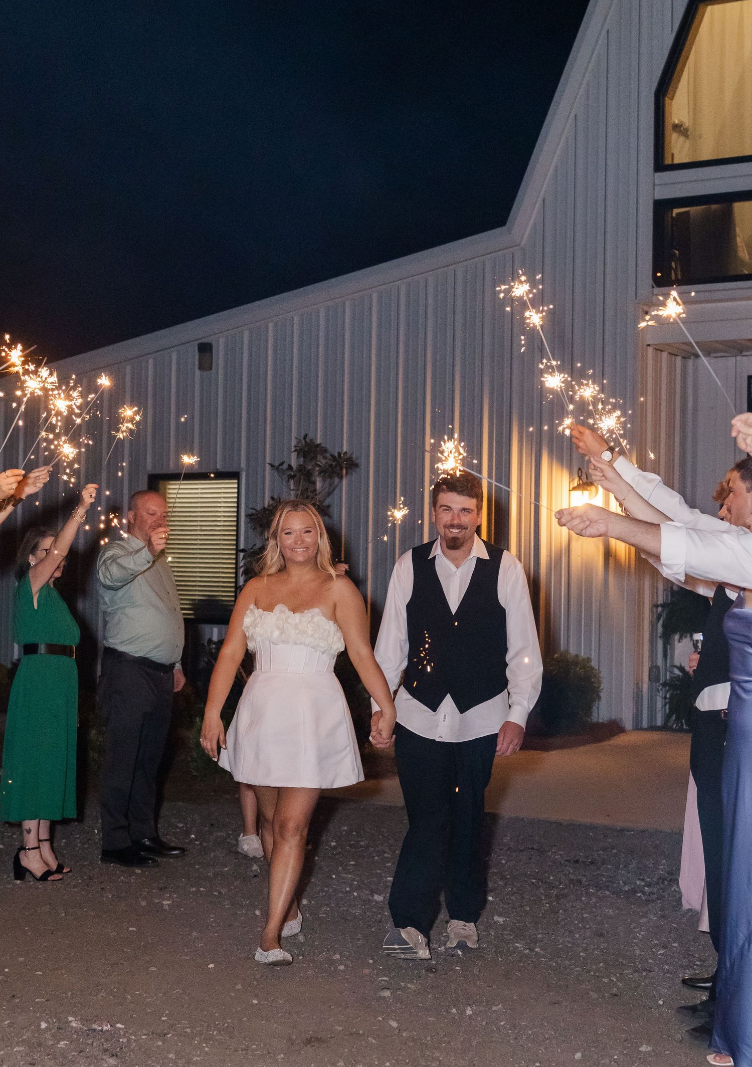 Newlyweds exit wedding venue through sparkler tunnel at night holding hands.