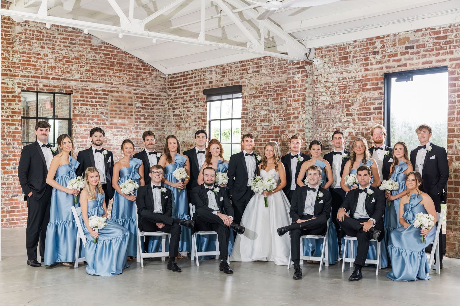 Large wedding party in blue gowns and black tuxedos posing in rustic brick venue with white ceilings.