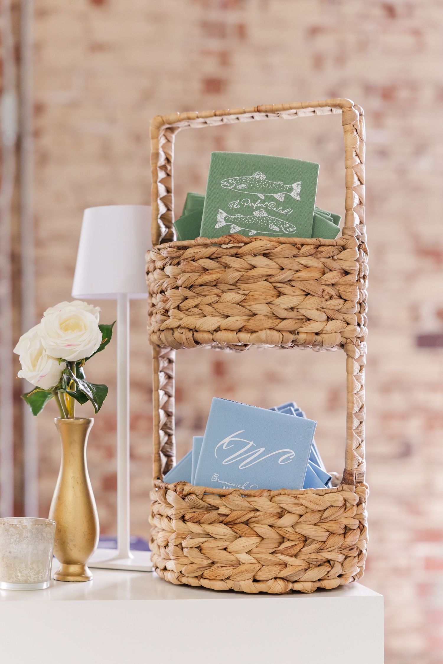 Woven basket organizer holding green and blue wedding programs beside a gold vase with white roses.