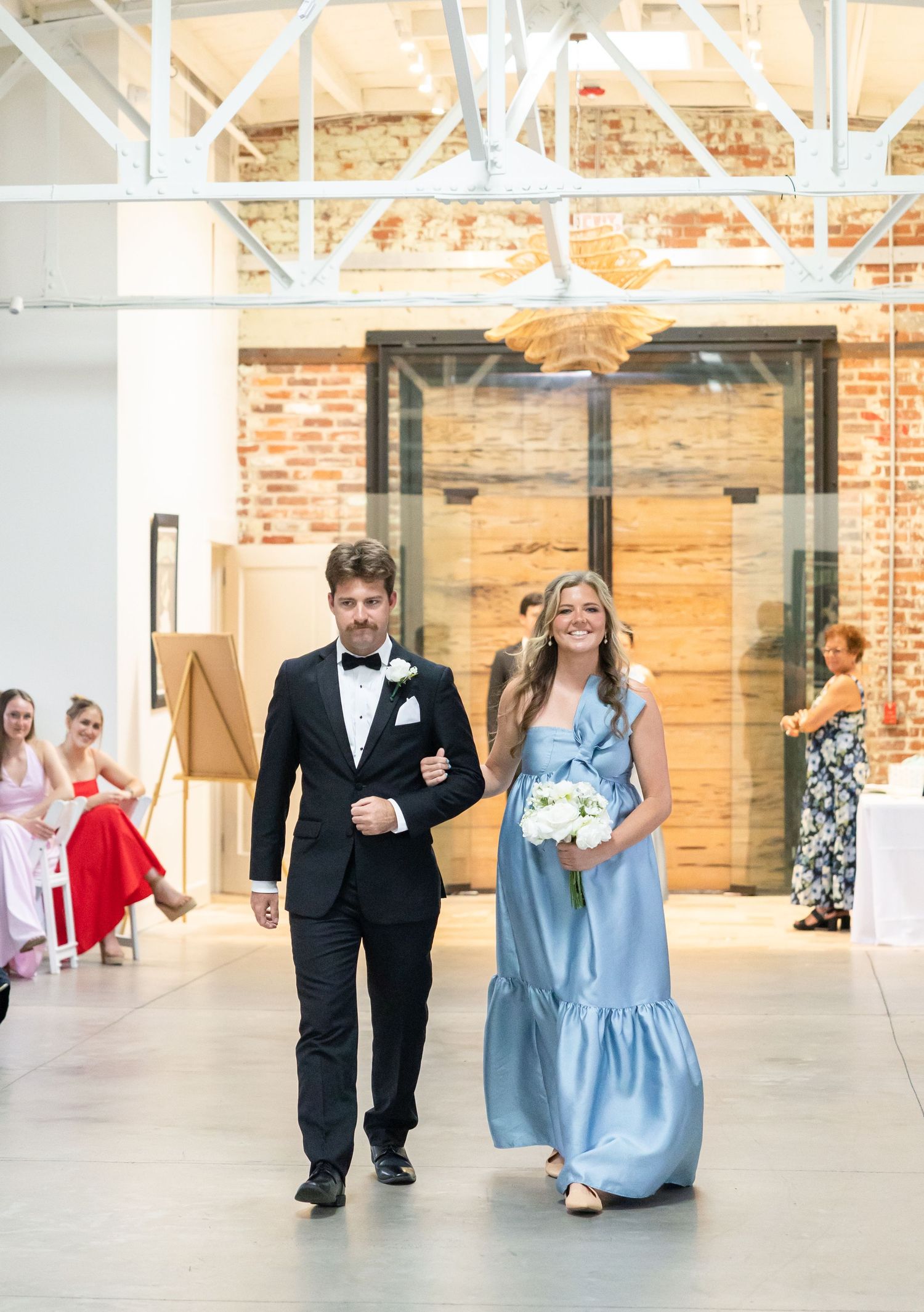 Bridesmaid in blue satin gown walks down aisle with groomsman in black tuxedo at wedding ceremony.