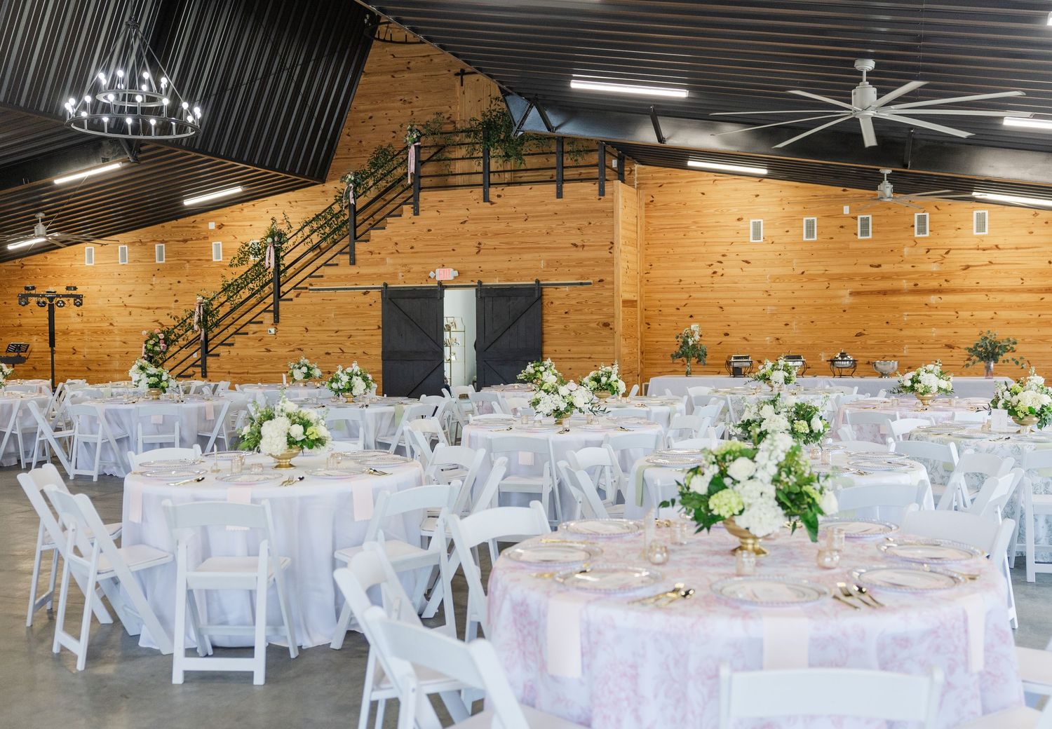 Rustic barn wedding reception venue with white chairs, round tables, floral centerpieces, and wooden walls.
