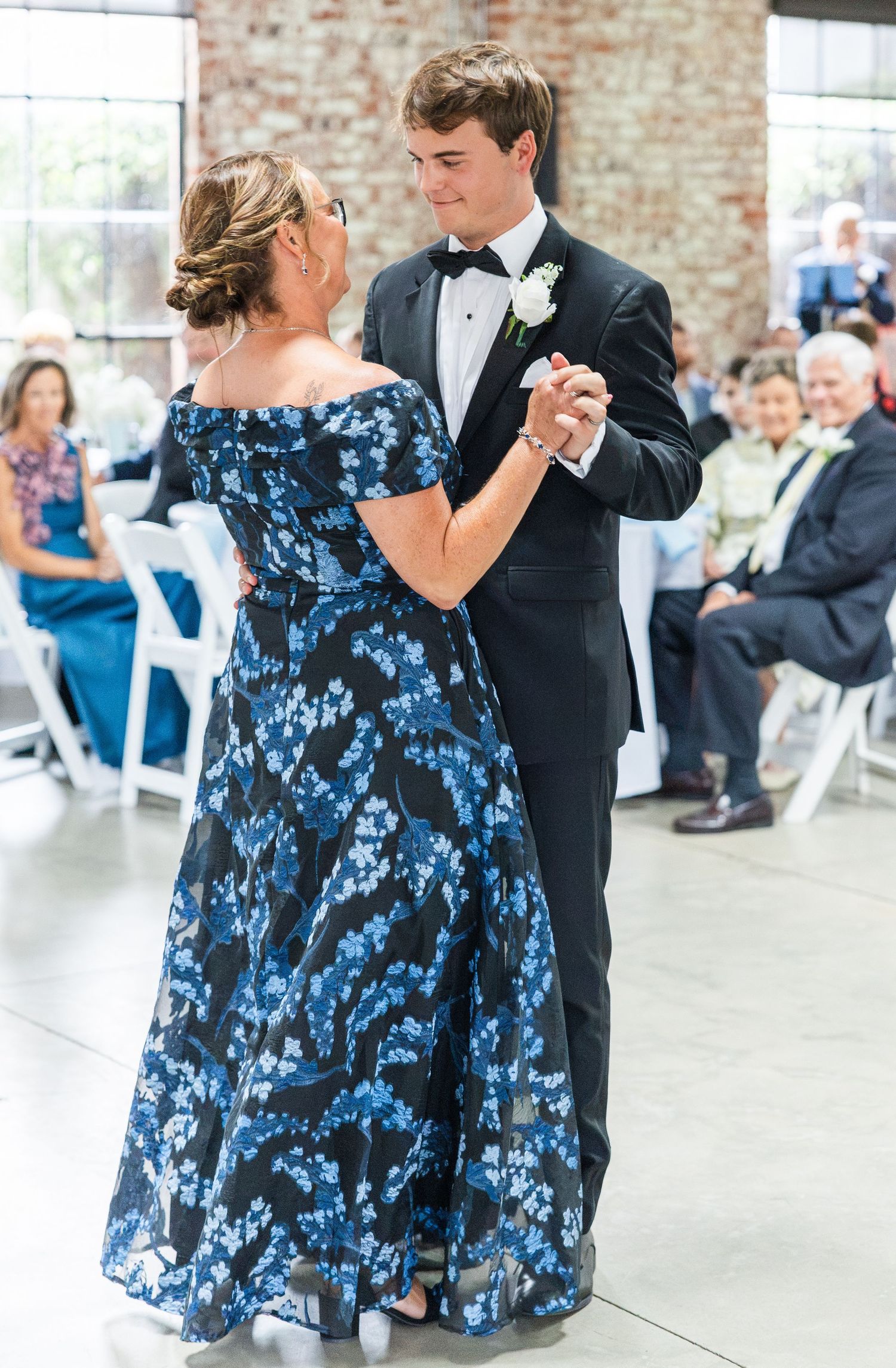 Groom in black tuxedo sharing a tender mother-son dance at wedding reception.
