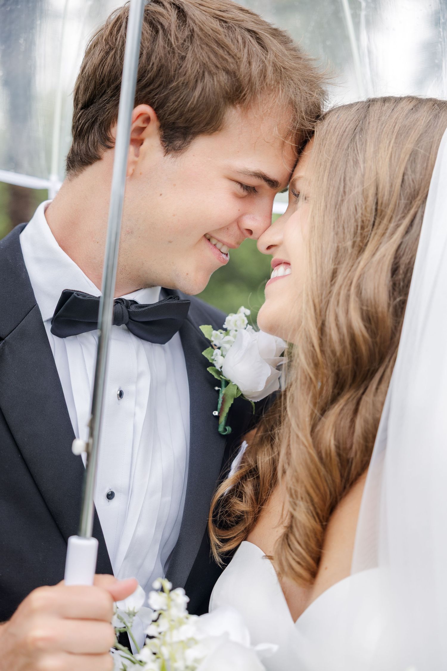 Smiling bride and groom share a romantic moment under a clear umbrella on their wedding day.
