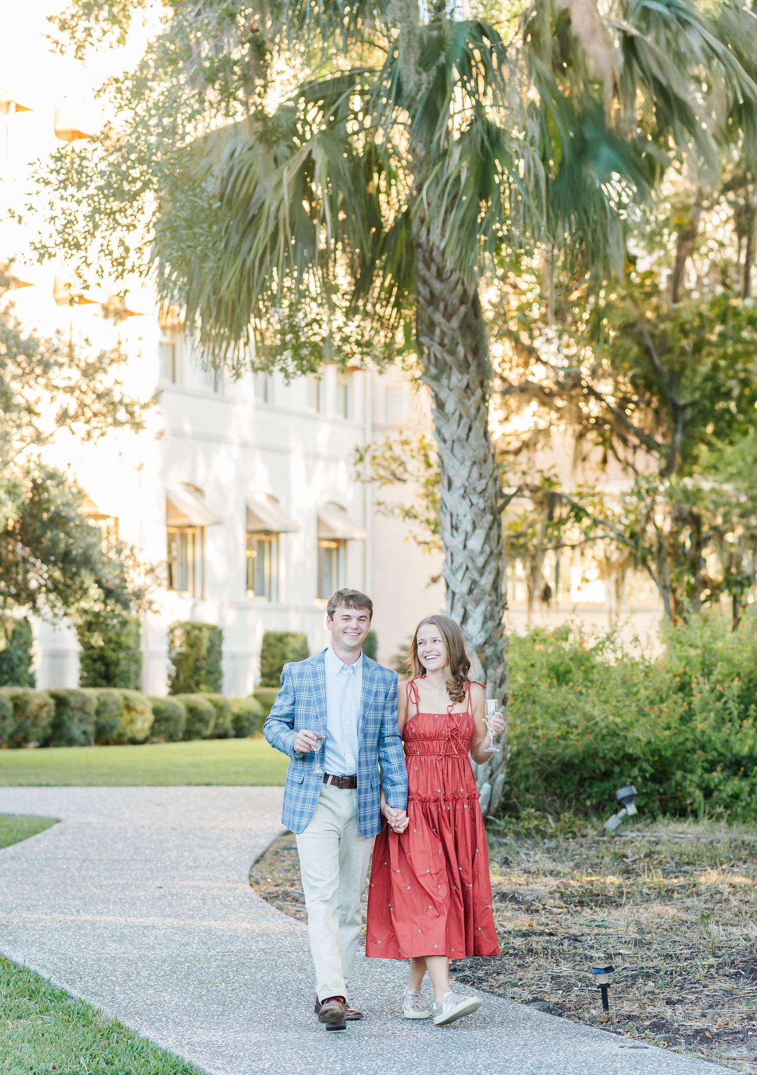 A couple strolls along a palm-lined pathway near a Mediterranean-style building.