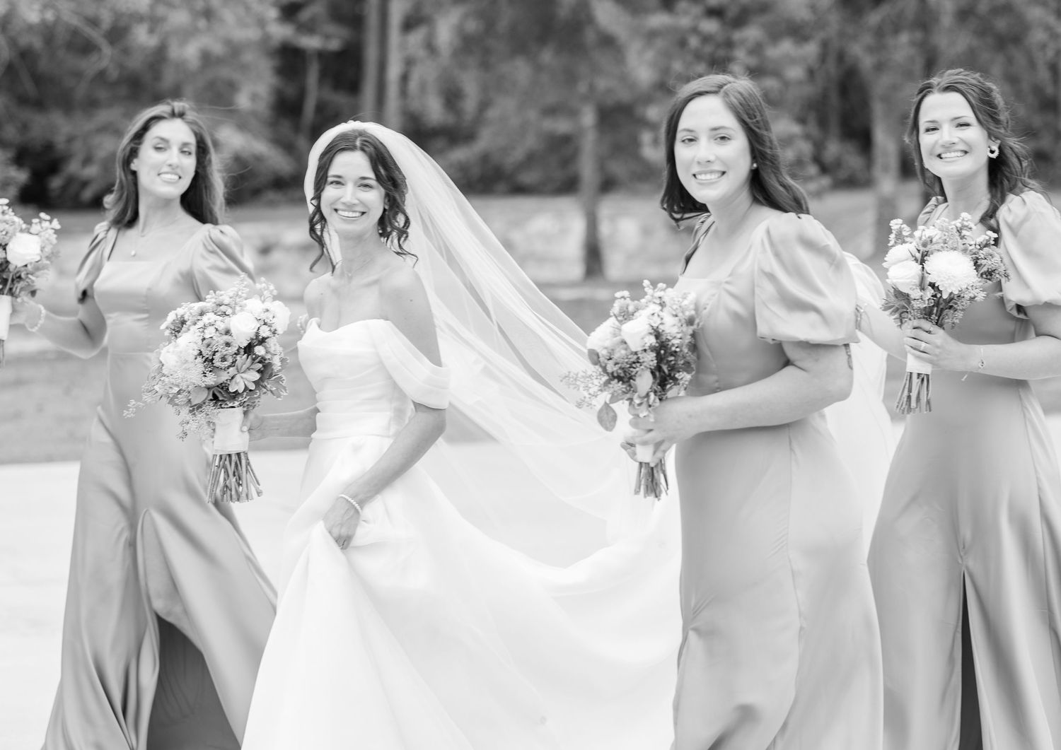 Joyful wedding party members in matching grey dresses laugh and dance together outdoors.