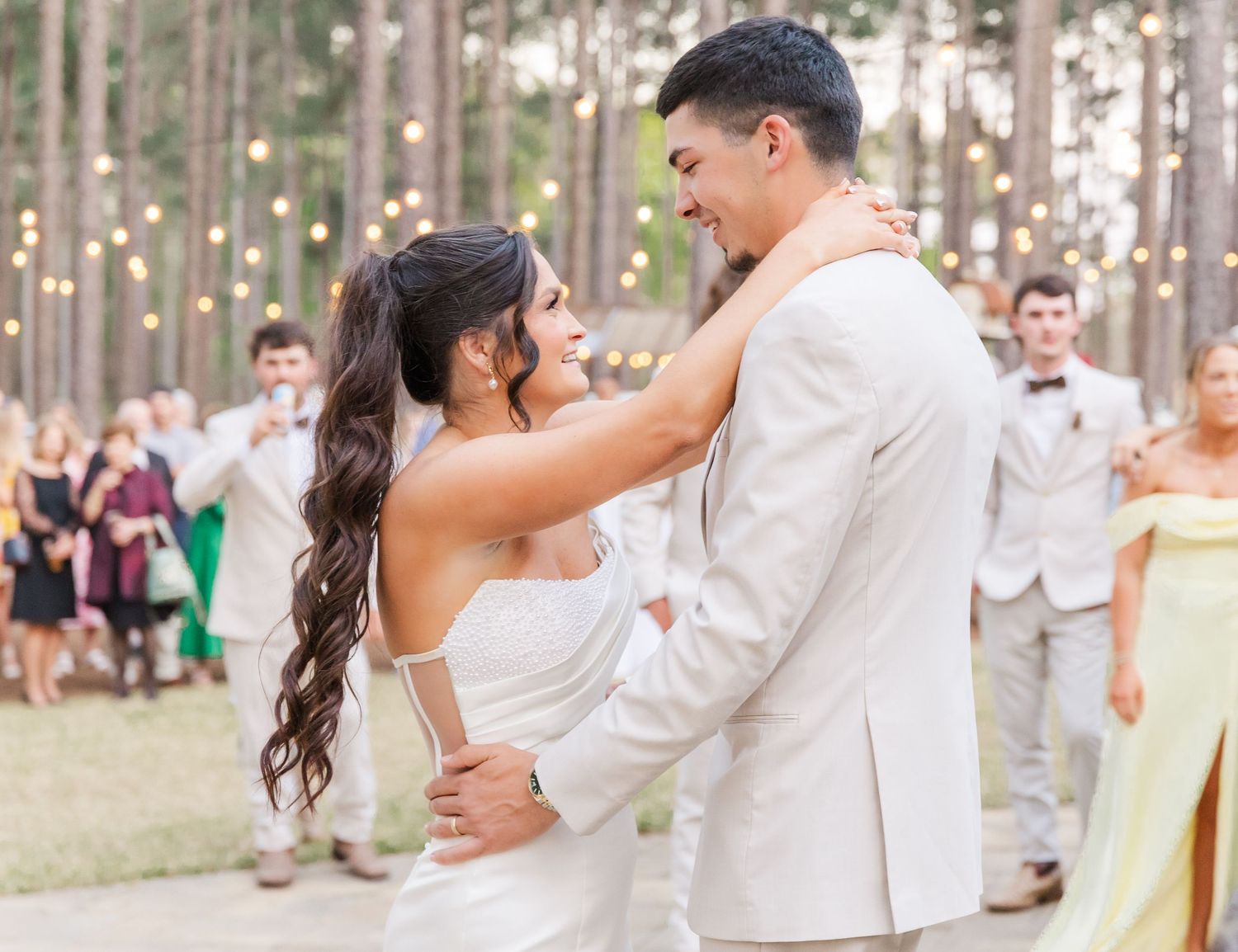 Bride and groom smiling during first dance outdoors with string lights and wedding guests in background.