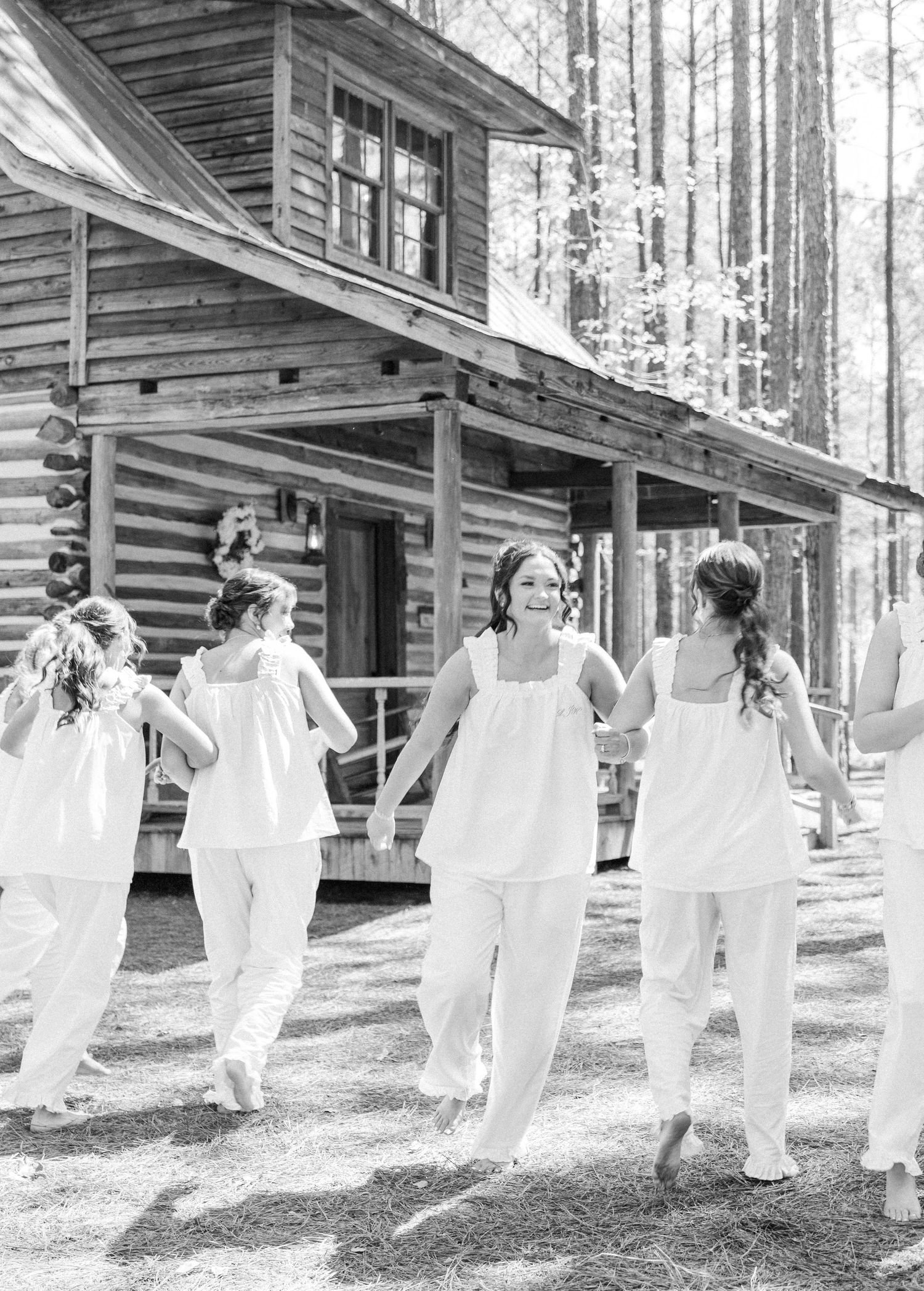 Bridesmaids in white outfits walking and laughing outside a rustic log cabin wedding venue.