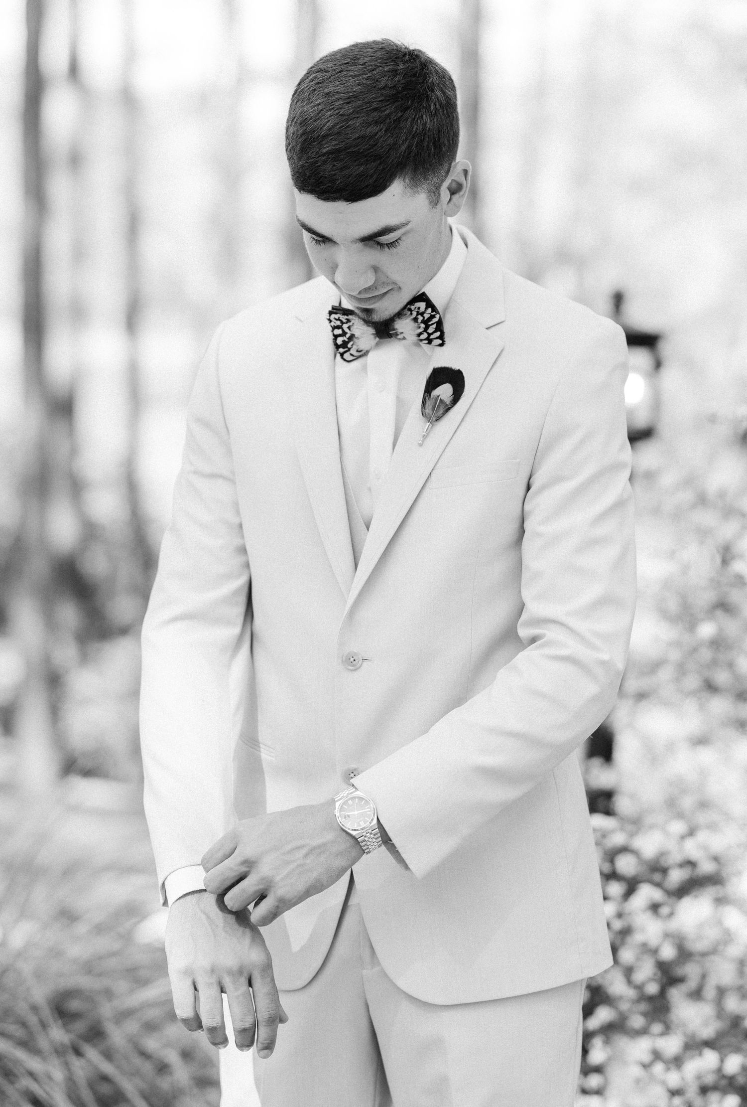 Elegant groom in cream suit adjusting cufflinks with a patterned bow tie in a black and white outdoor portrait.