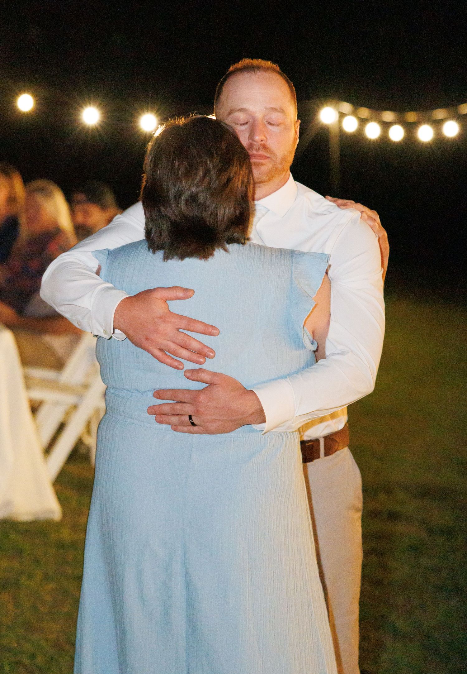 Groom in white shirt sharing a tender embrace with a guest in light blue dress at night reception.