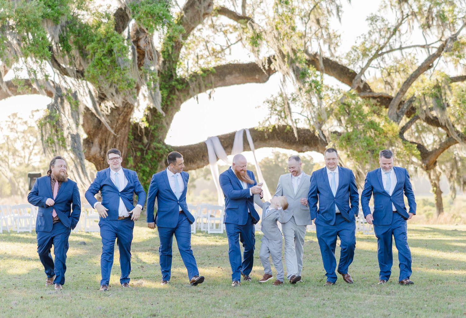 Groom in gray suit walks with groomsmen in navy suits under Spanish moss-draped oak trees at outdoor wedding.