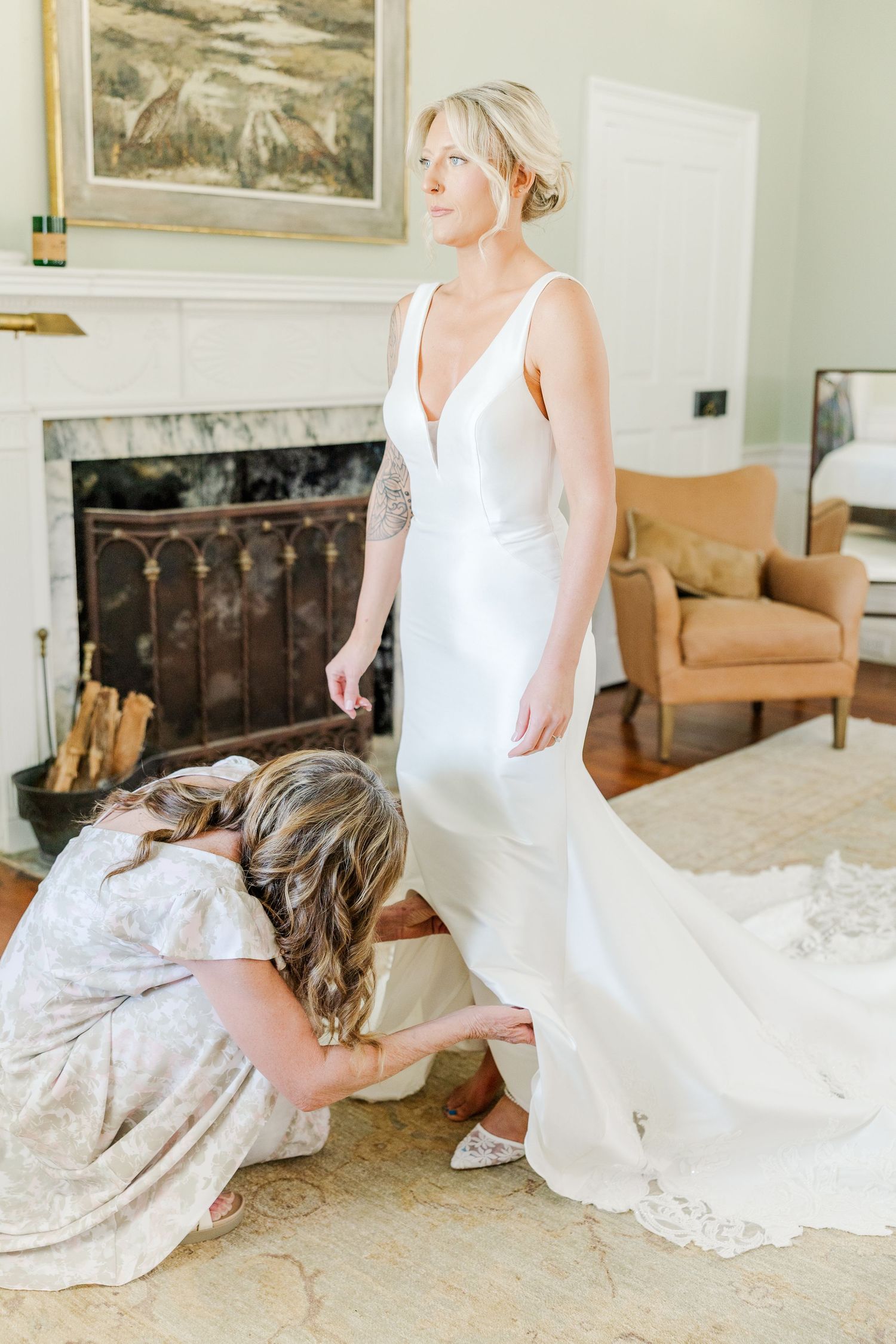 Bridesmaid helps bride with shoes as she stands in elegant white gown near fireplace indoors.