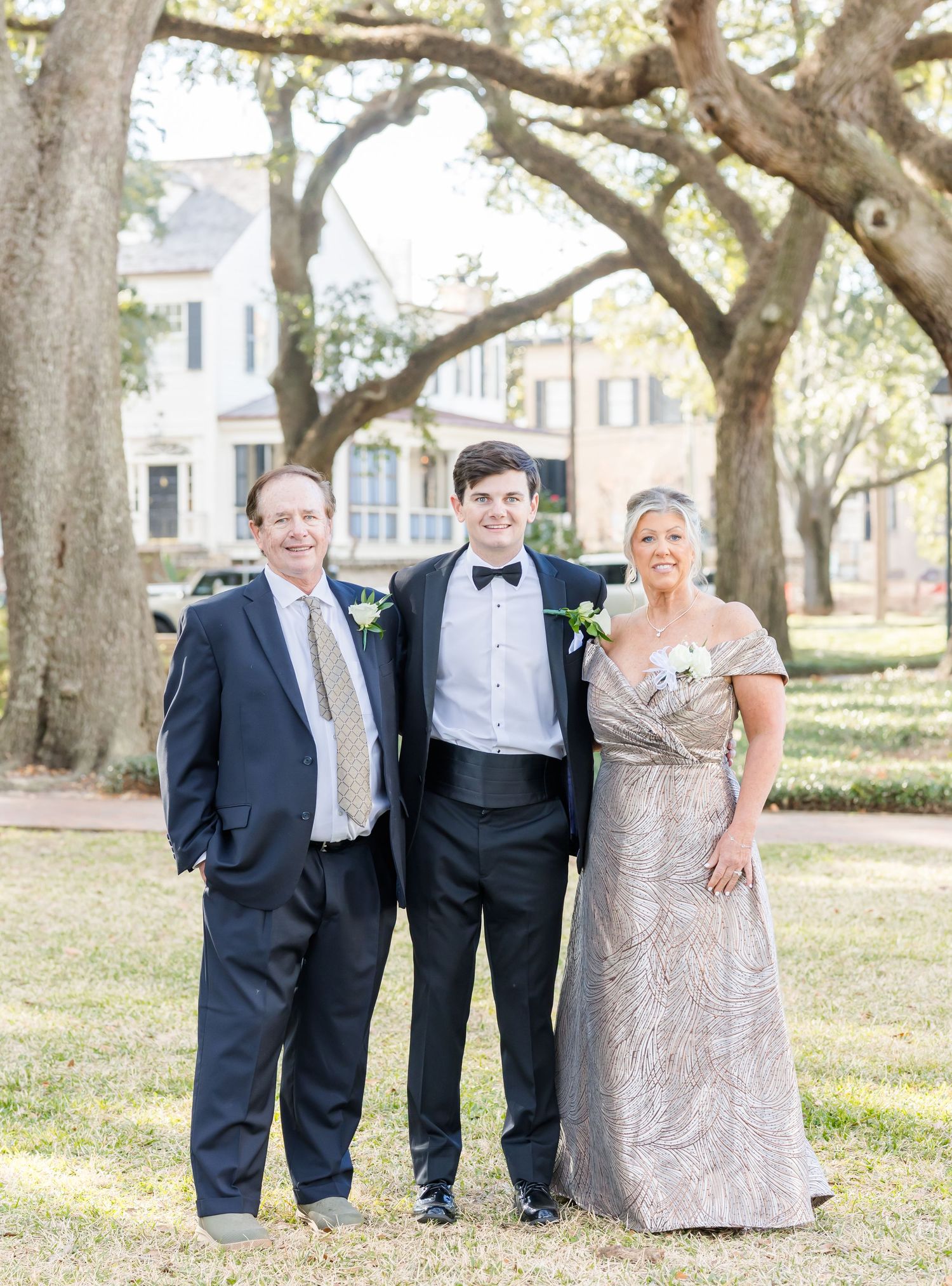 Wedding party poses under grand oak trees wearing formal attire in a garden setting.