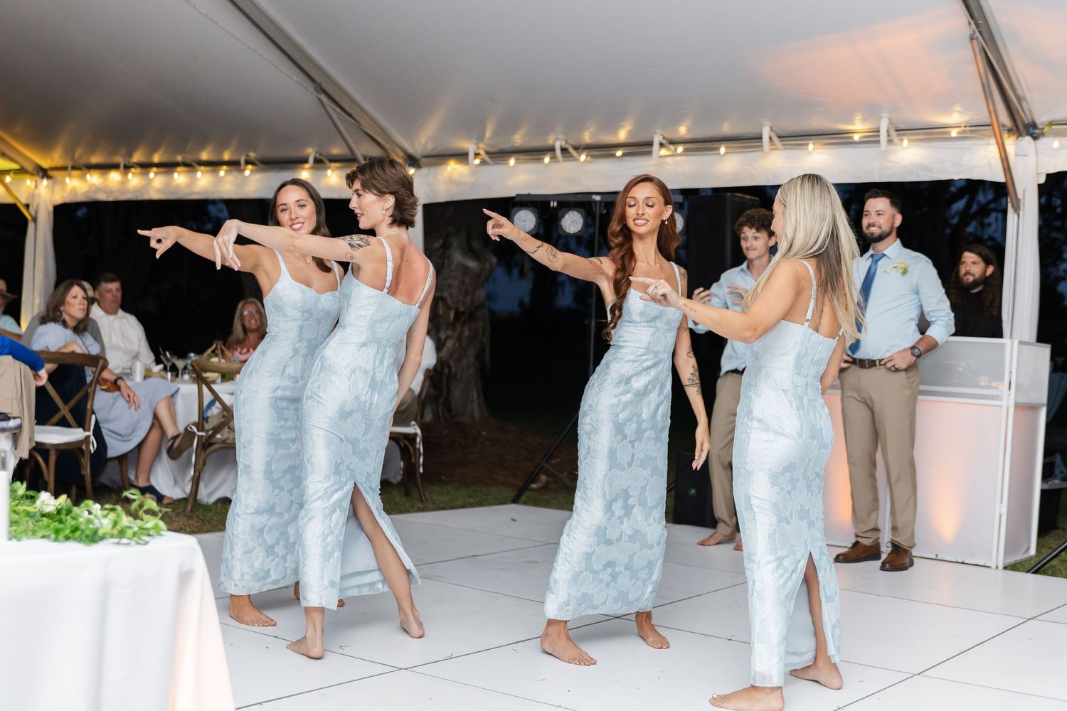 Wedding party members in light blue dresses dance in a line at an outdoor reception.