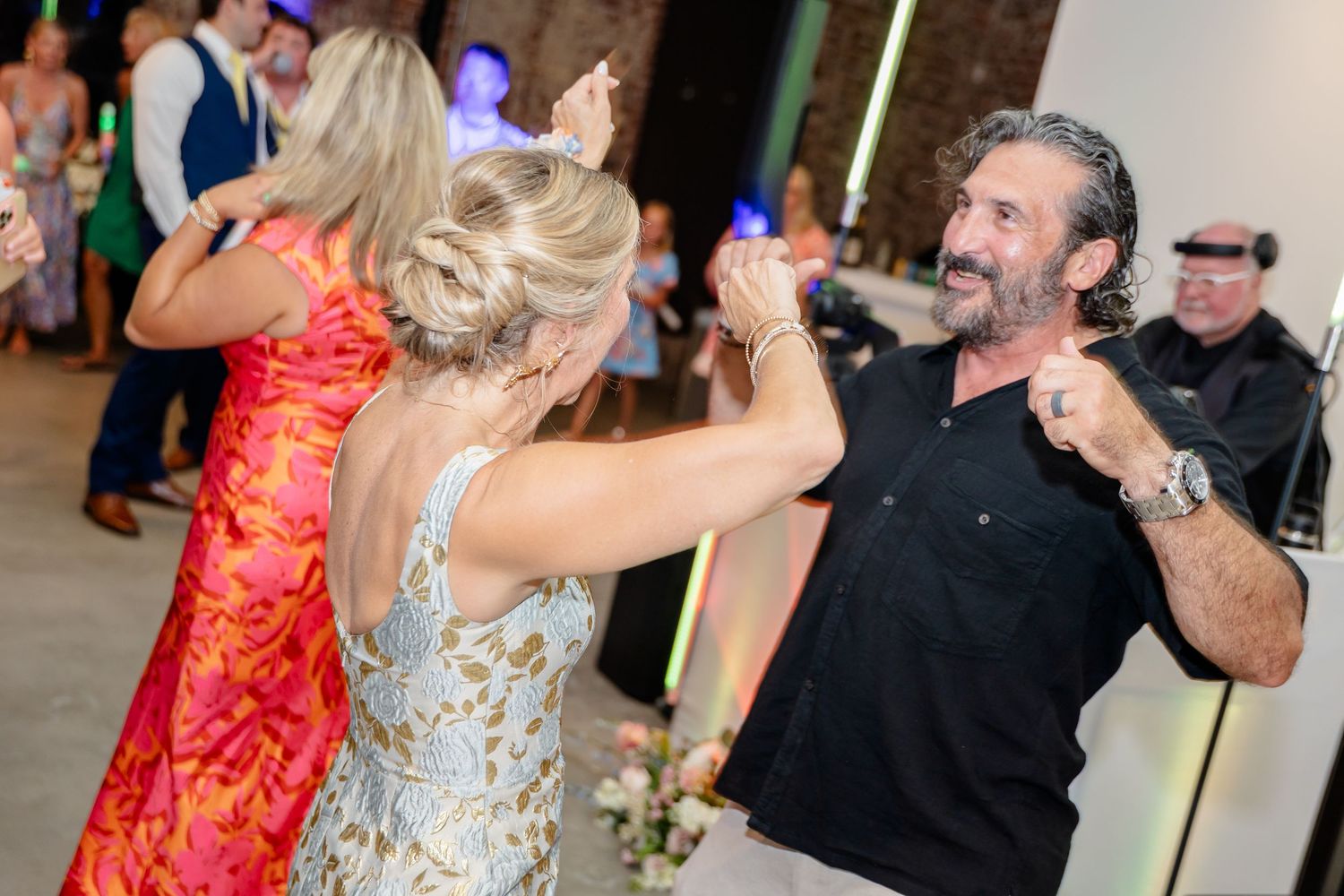 Party guests dancing with light-up foam sticks at a wedding reception in a brick-walled venue.