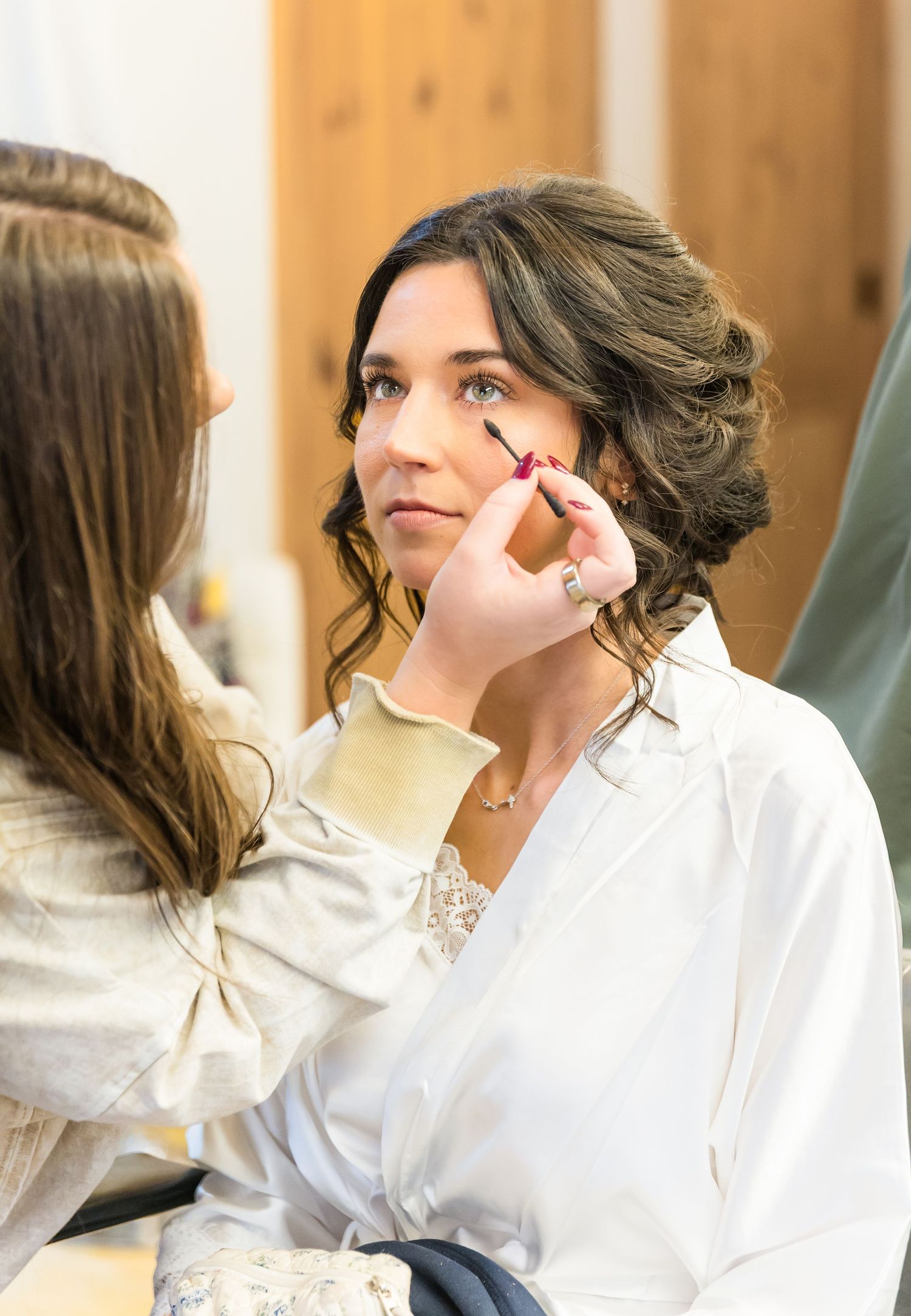 Professional makeup artist applies cosmetics to a client wearing a white shirt in a sunlit room.