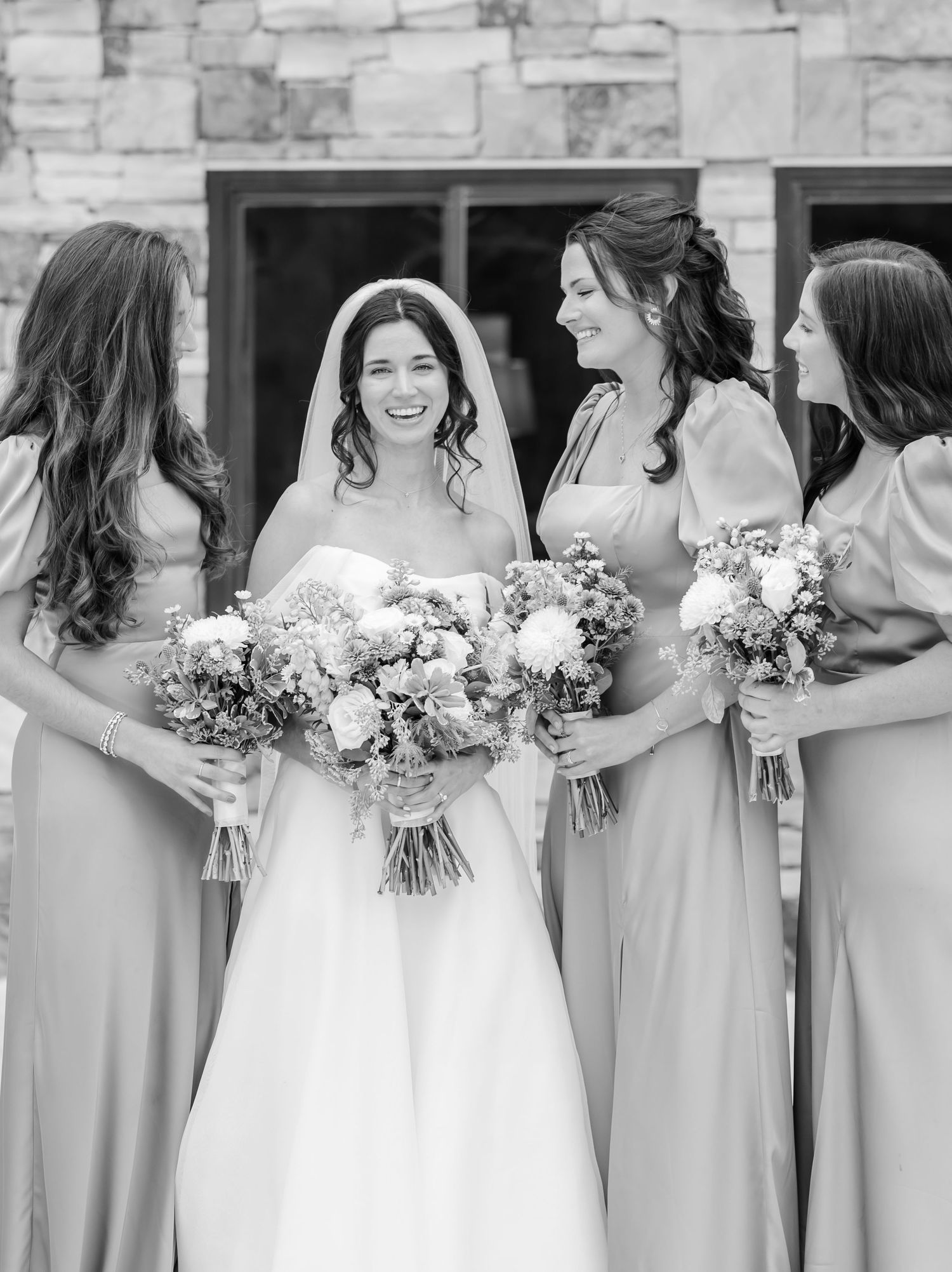 Wedding party wearing light blue bridesmaid dresses and white gowns holding floral bouquets against stone wall backdrop.