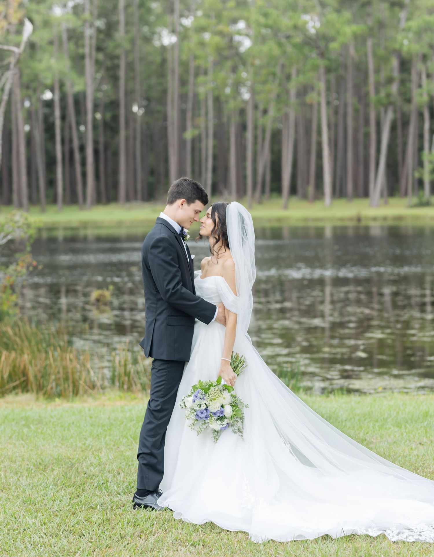 Elegant wedding photos by lakeside with long flowing veil and natural woodland backdrop.