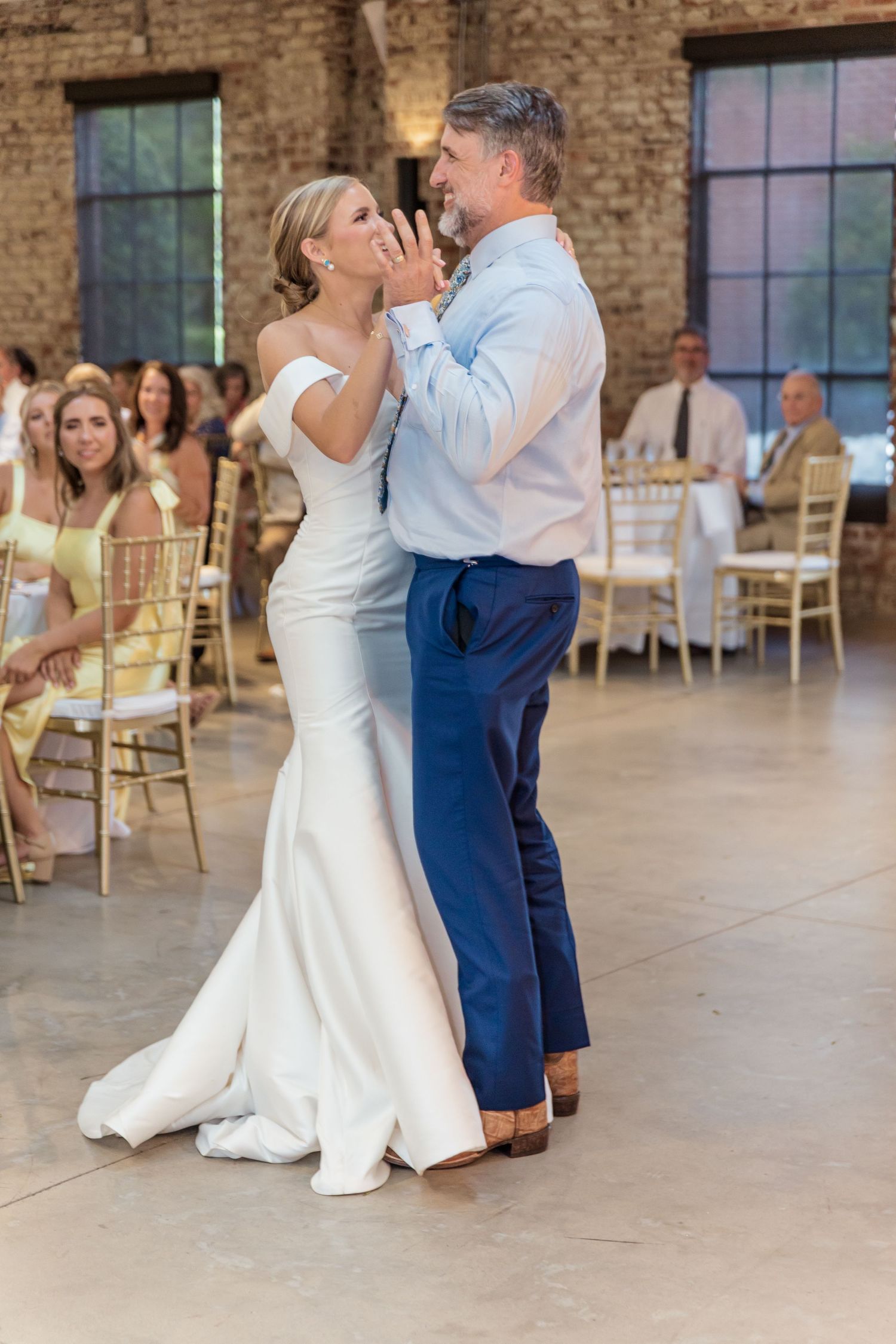 A couple shares their first dance at a wedding reception in an industrial-style venue with exposed brick walls and gold chairs.