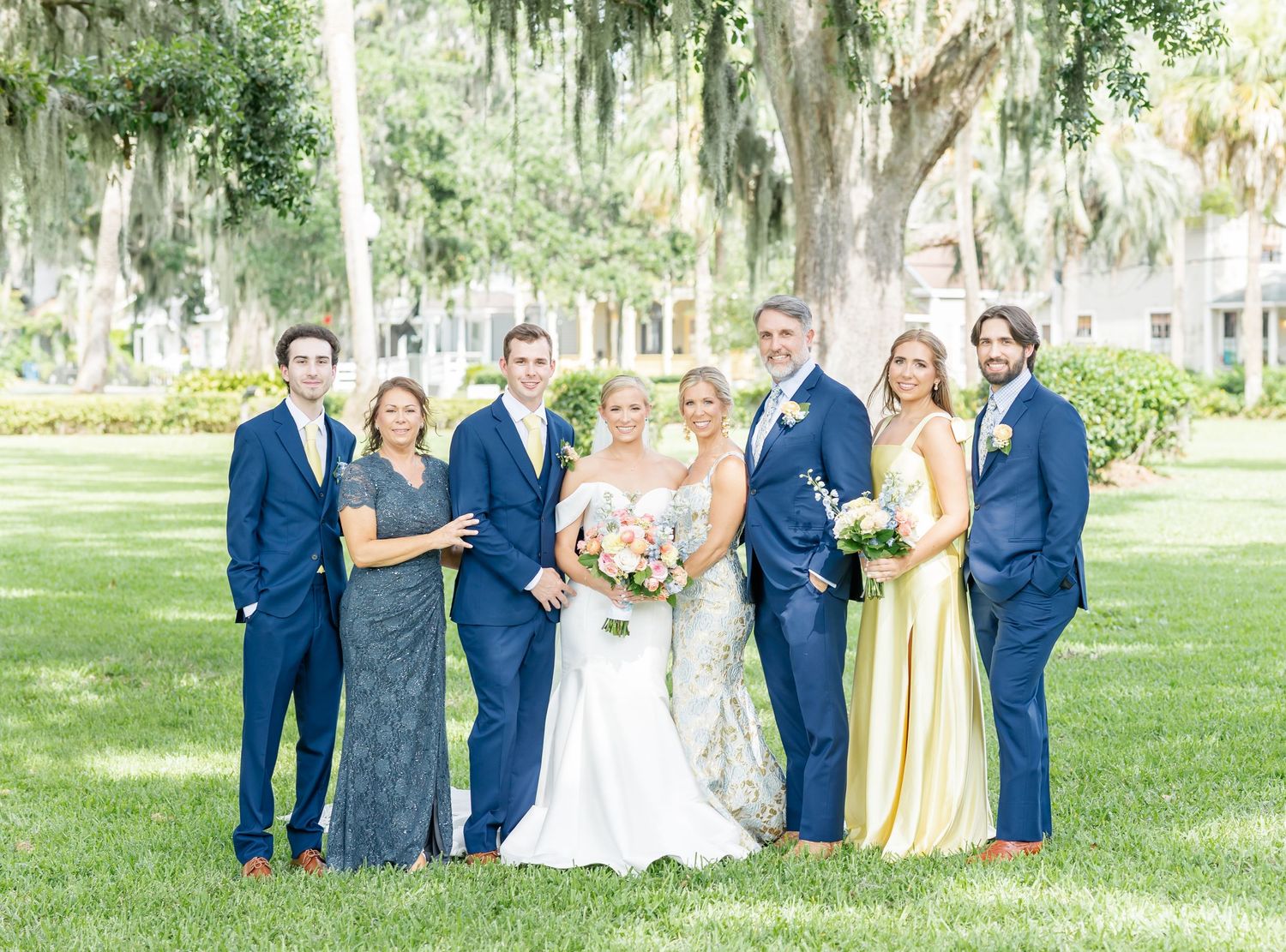 Wedding party in navy suits and yellow dresses poses together on a lush green lawn under Spanish moss trees.