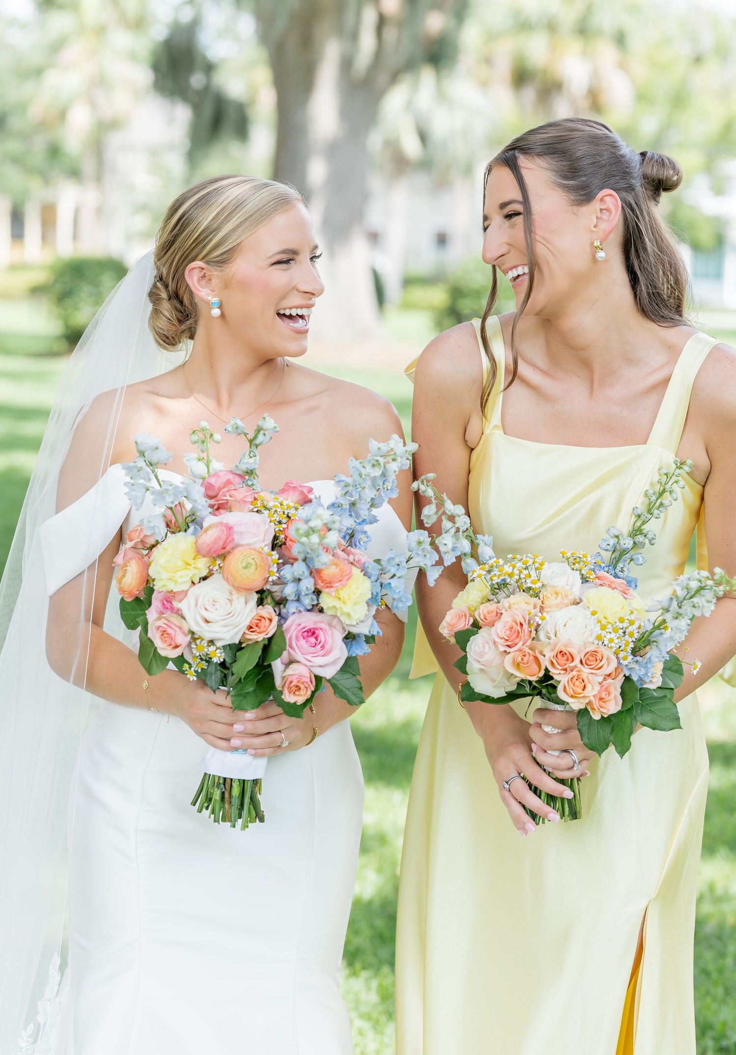 Wedding party members holding vibrant peach and pink floral bouquets while laughing together outdoors.