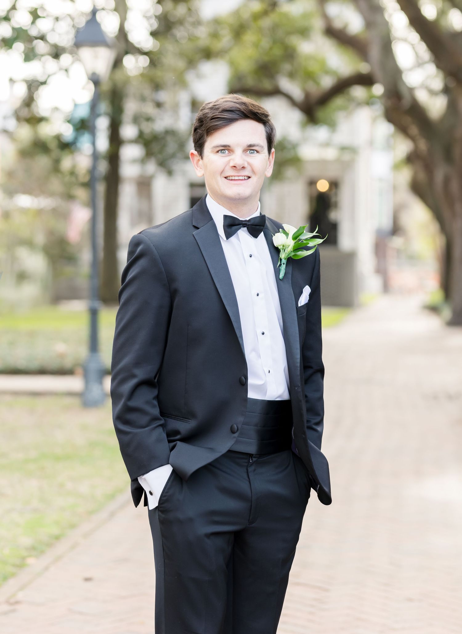 Groom in classic black tuxedo poses confidently on tree-lined pathway during wedding portraits.
