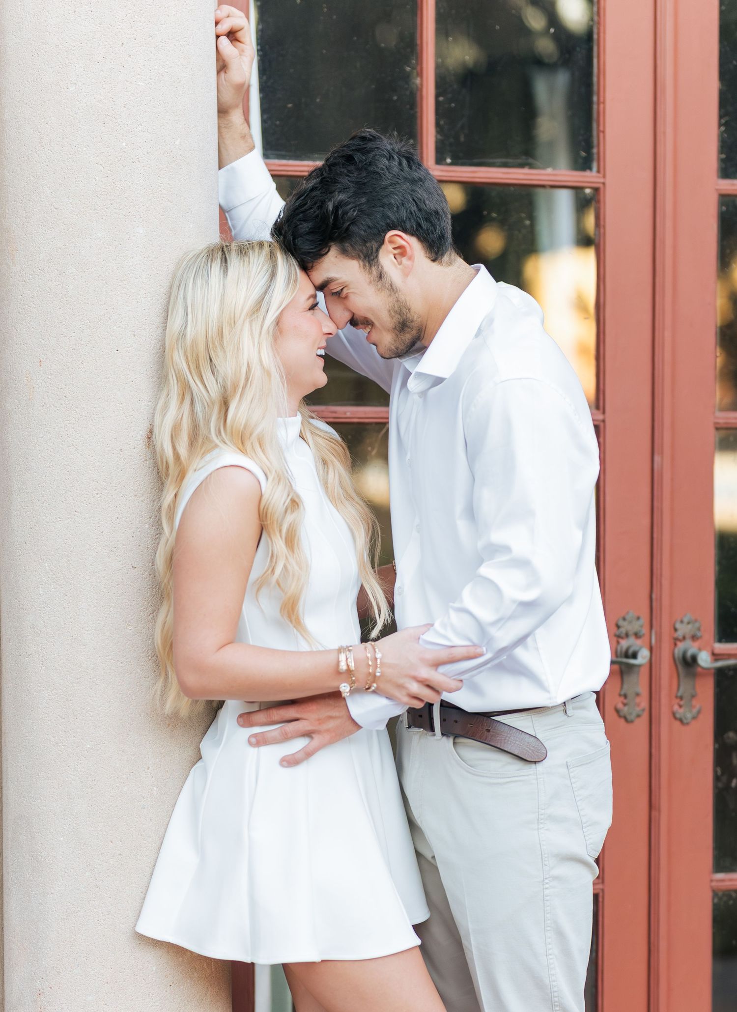 A romantic engagement photo session showing a couple in white and grey outfits posing against a rustic doorway.