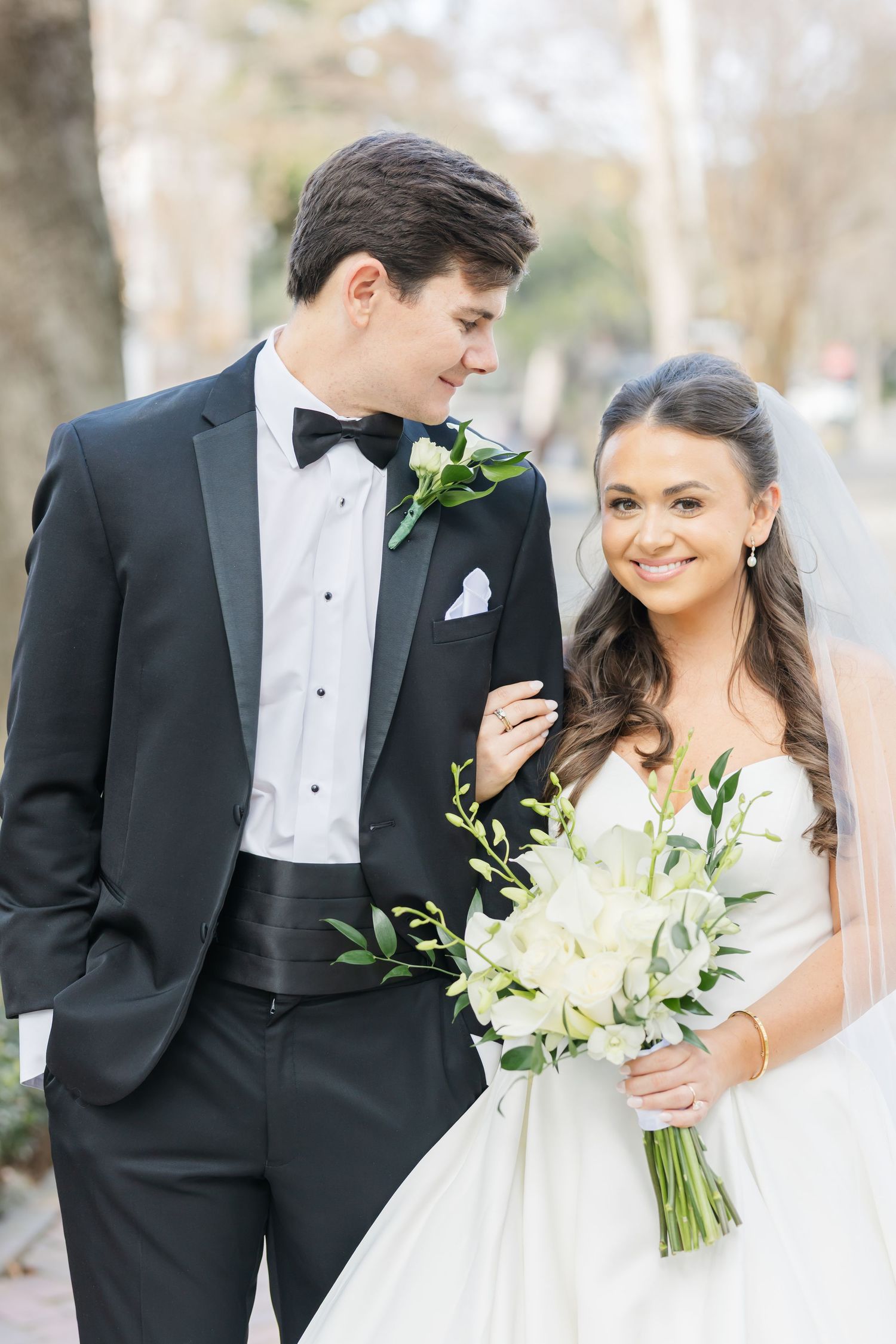 A couple in formal wedding attire pose together outdoors with white floral bouquet and boutonniere.