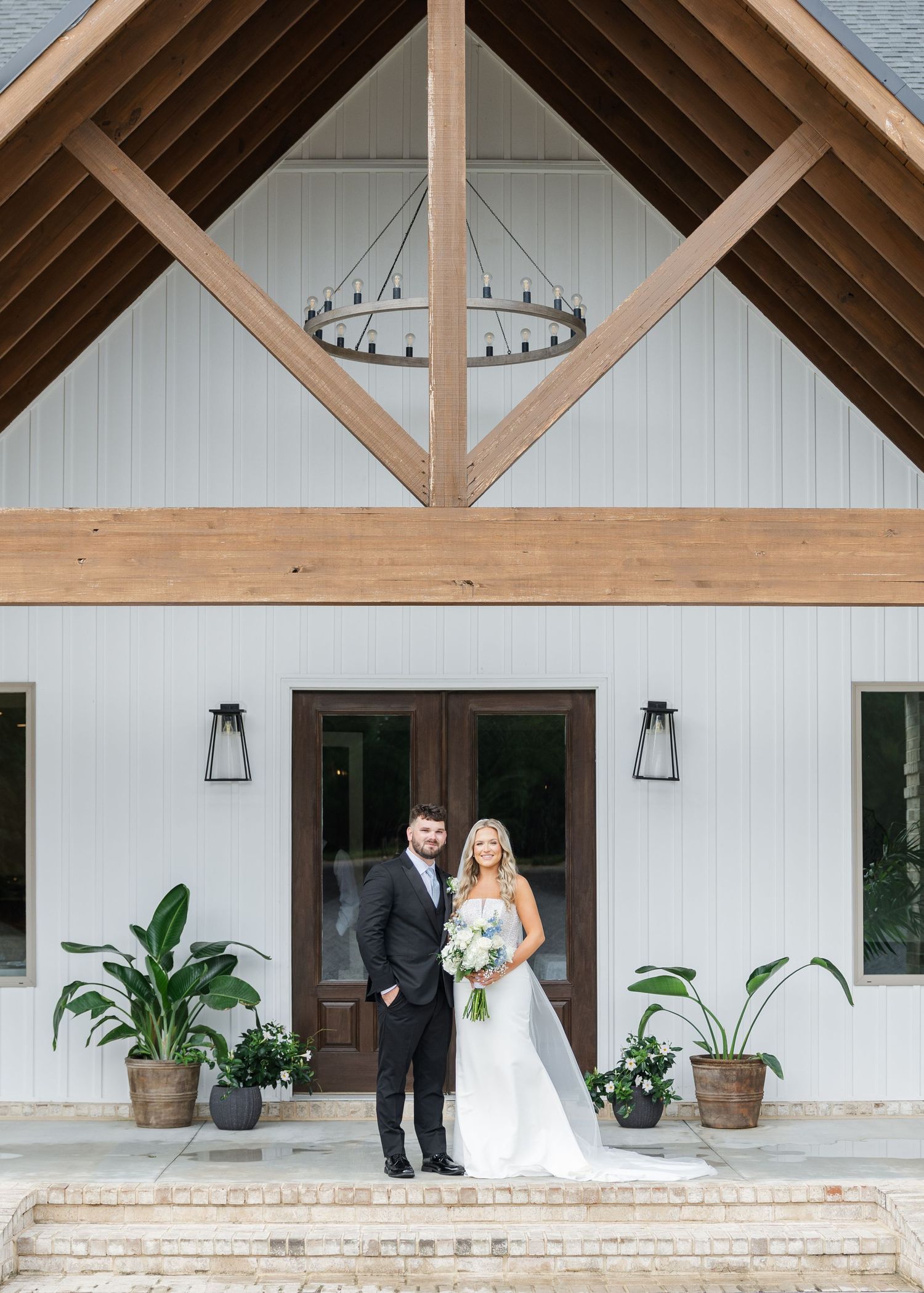 Bride and groom posing at entrance of white barn wedding venue with wood beam accents and lush greenery.