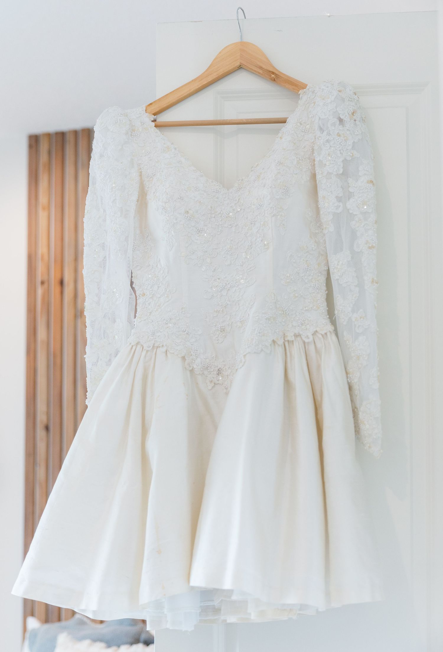 Short lace bridal gown with long sleeves and full skirt hanging on wooden hanger.