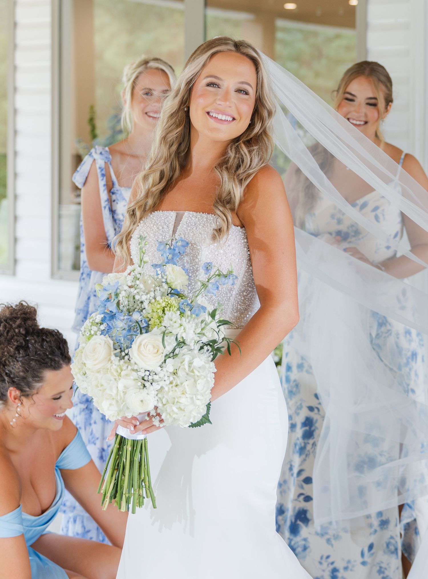 Radiant bride in beaded gown holding blue and white bouquet surrounded by bridesmaids in floral dresses.