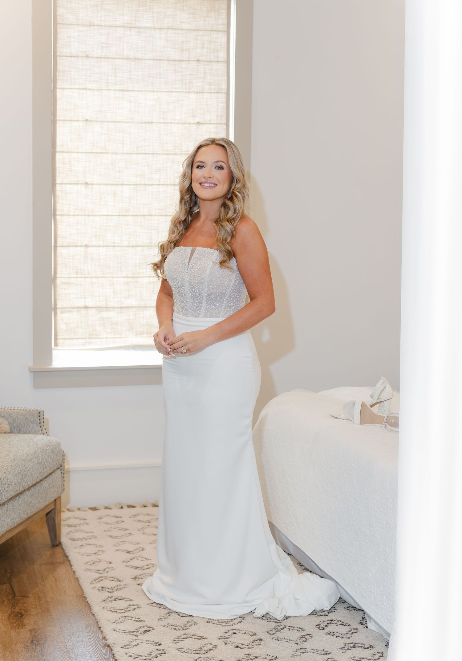 Bride in strapless fitted white wedding gown standing by window in bridal suite.
