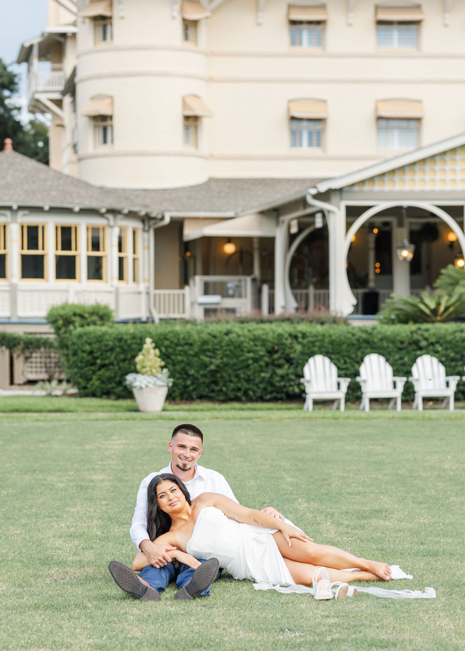 Couple relaxing on green lawn in front of elegant yellow historic hotel with white chairs.