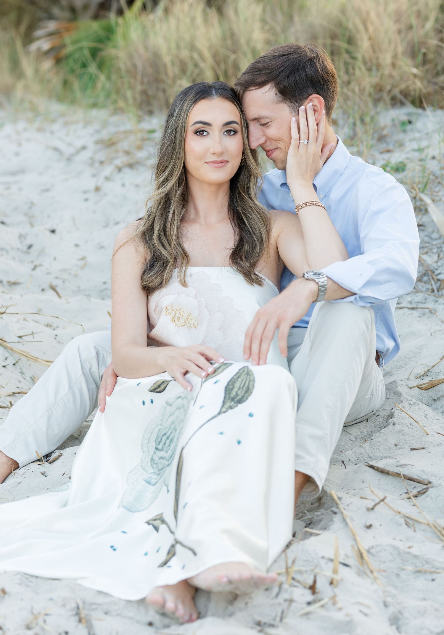 Couple sitting on sandy beach, woman in floral skirt leaning against man in blue shirt during romantic engagement photo session.