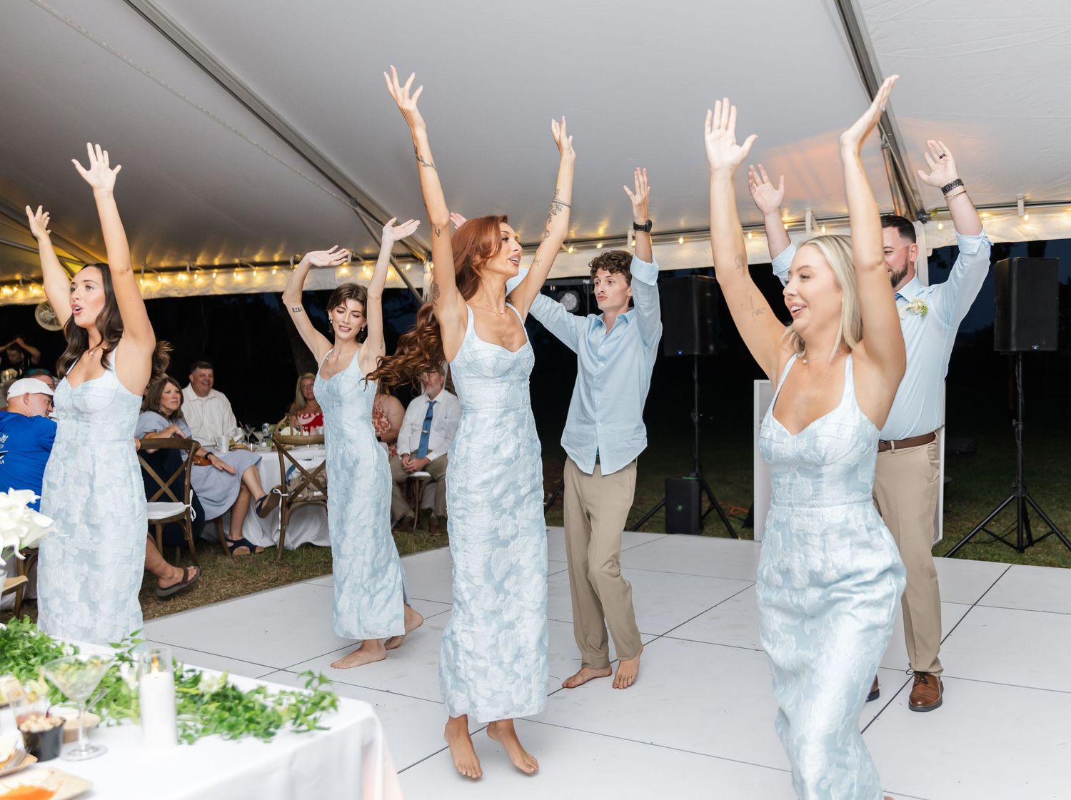 Wedding celebration with people dancing and raising their arms joyfully under a white tent.