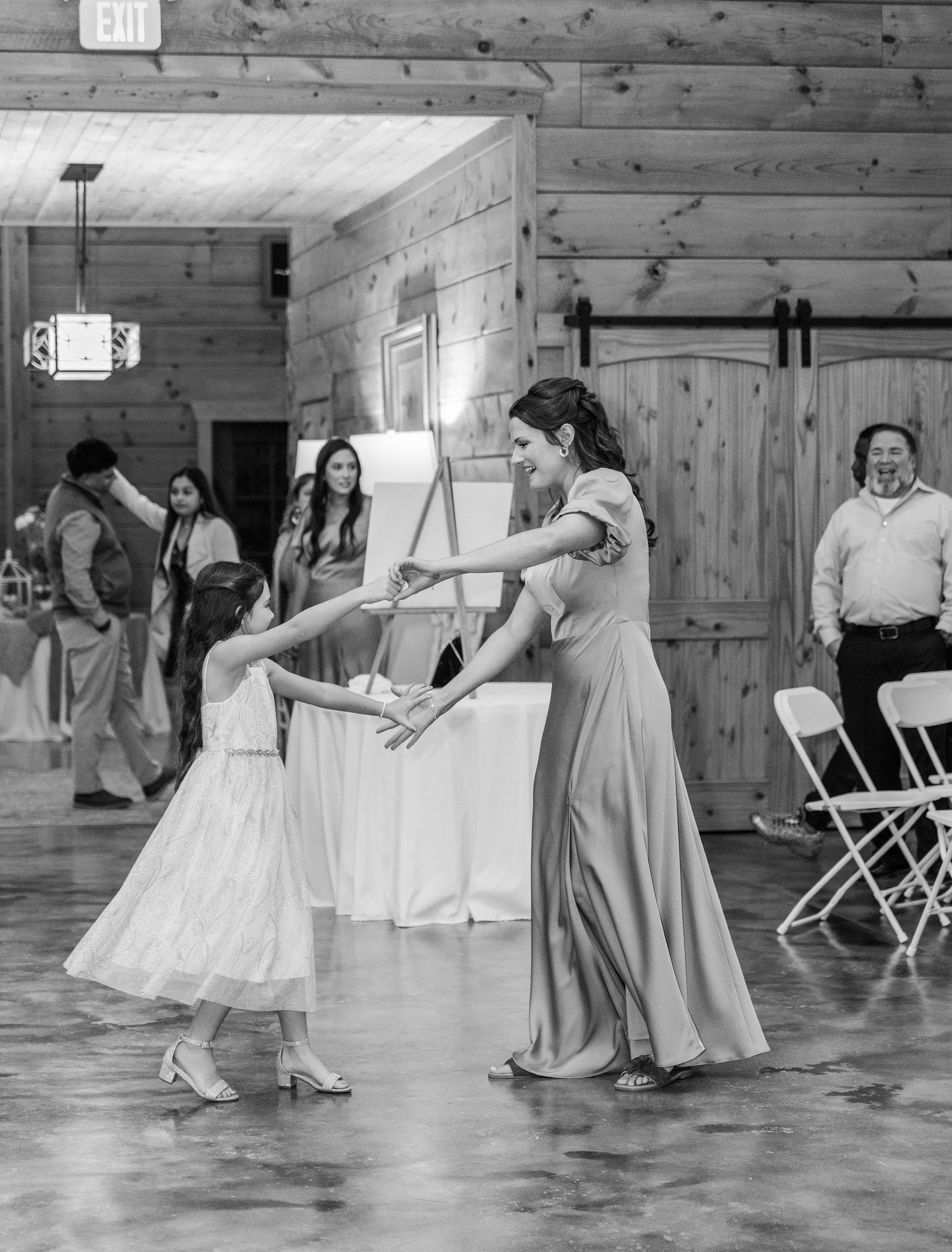 A black and white photo of people dancing together in a rustic wooden barn venue during a celebration.