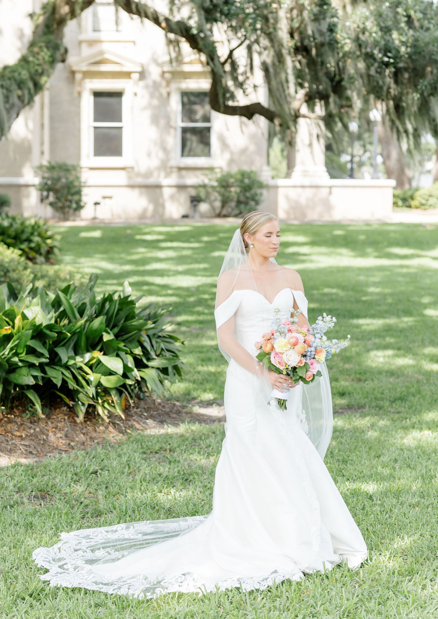A bride in a white lace wedding gown with a long train stands on a sunny lawn with Spanish moss draped trees in the background.