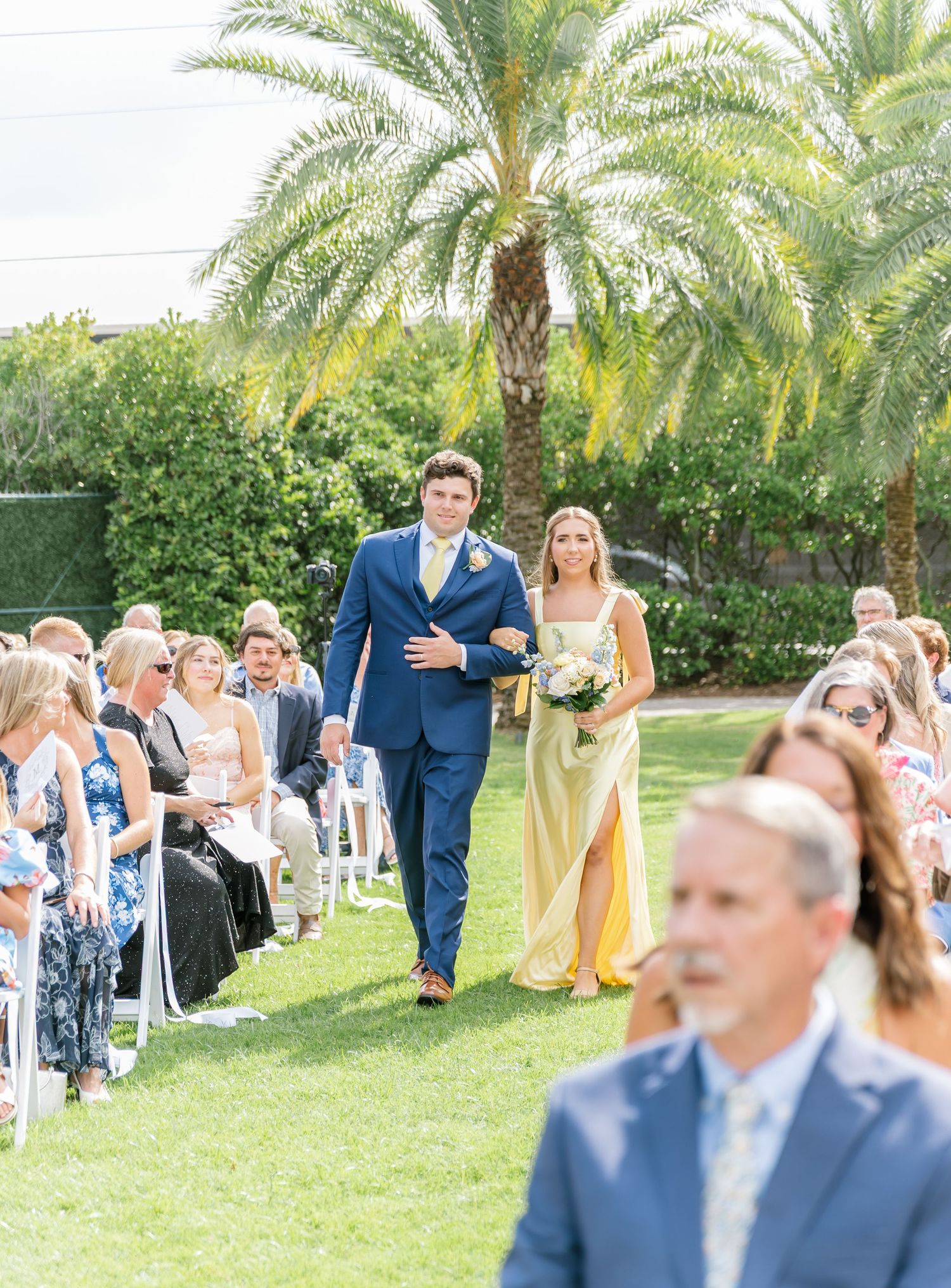 Wedding guests seated in white chairs on a lawn surrounded by tropical palm trees during an outdoor ceremony.