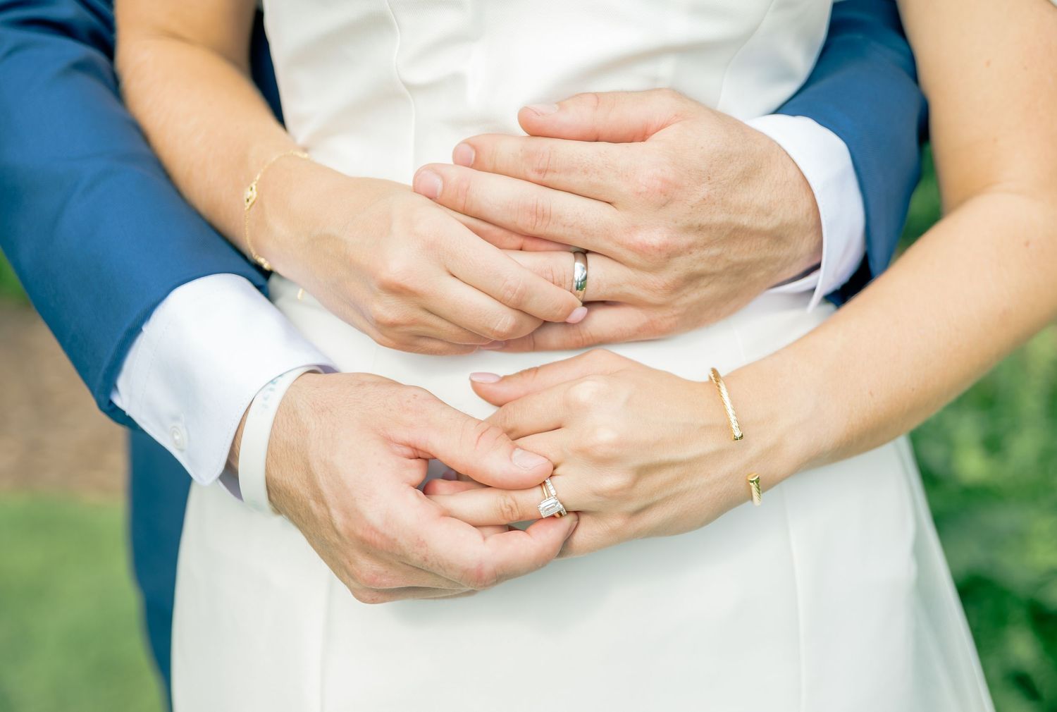 A row of hands clasped together in white wedding attire against a natural outdoor background.