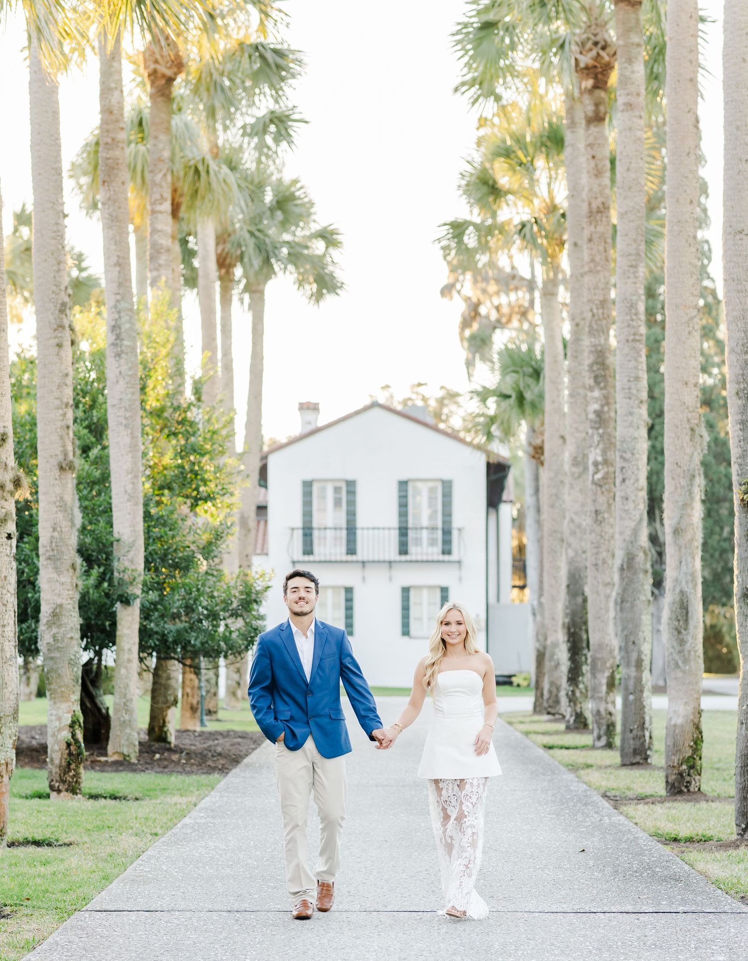 A pathway lined with majestic palm trees leads to a white building in a romantic outdoor setting.