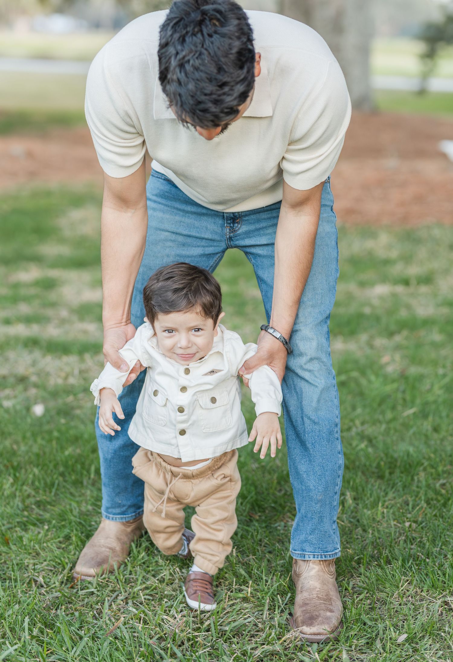 Parent helping small child take first steps on grassy lawn during outdoor photoshoot.