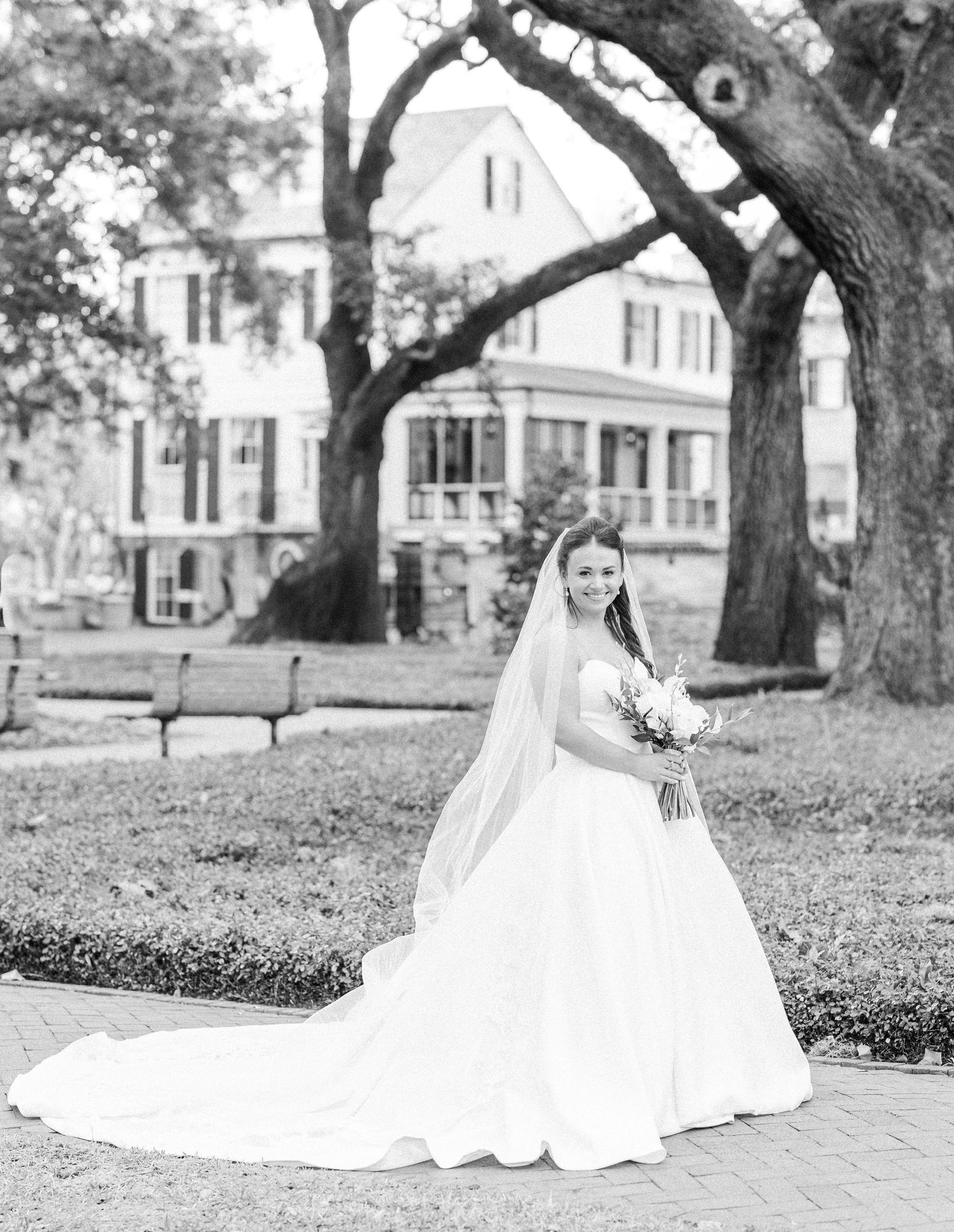 A bride in a flowing white gown sits gracefully on brick pavement under oak trees.