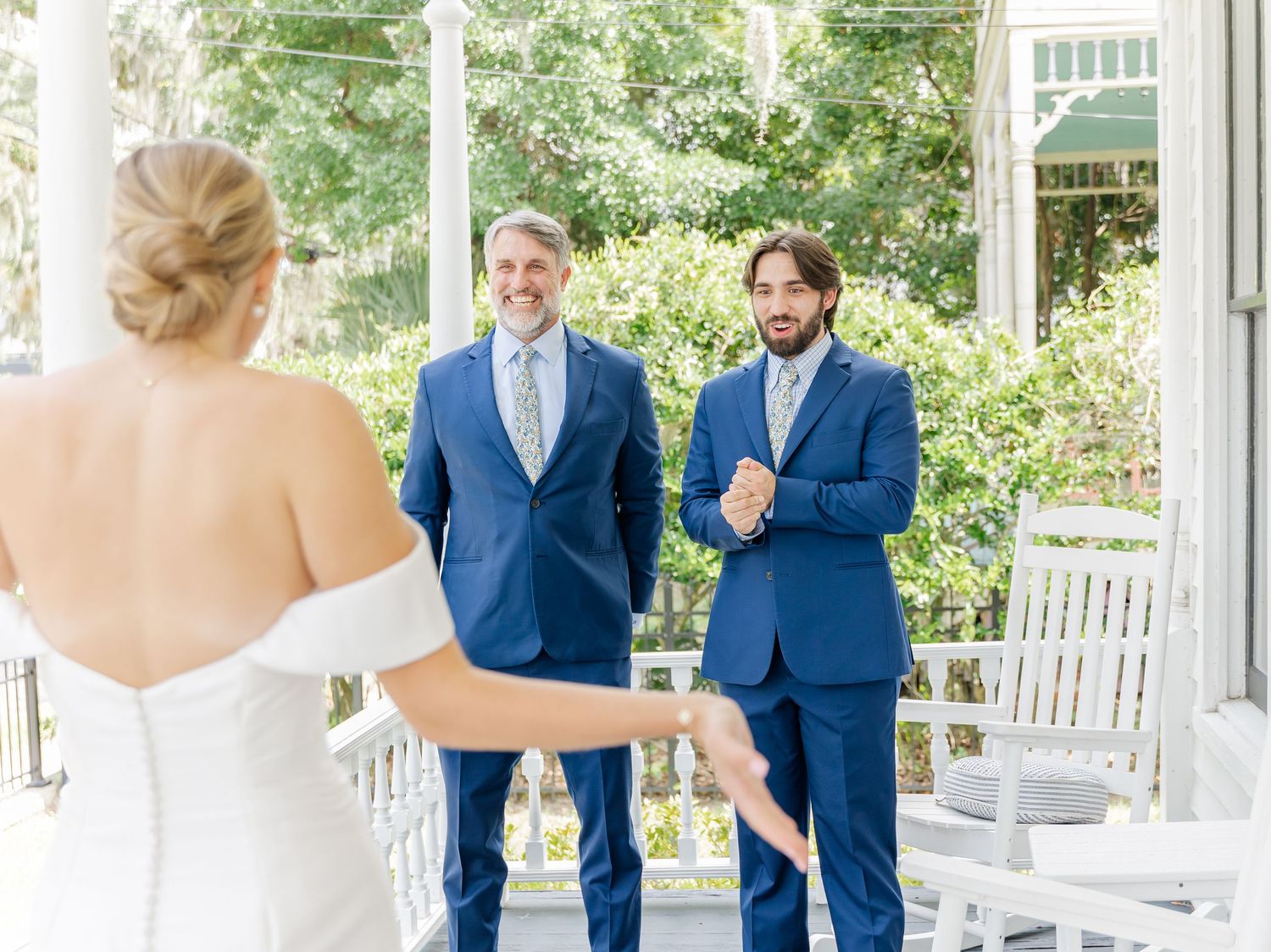 Wedding party members share joyful reactions during first look moment on a white porch surrounded by greenery.